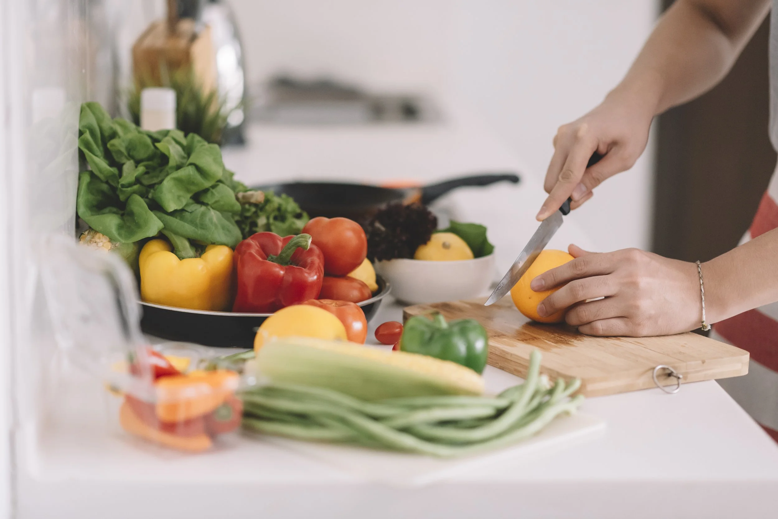 Person schneidet eine orangefarbene Paprika auf einem Schneidebrett in der Küche, umgeben von verschiedenen bunten Paprika, Tomaten, Frühlingszwiebeln und Salatblättern.