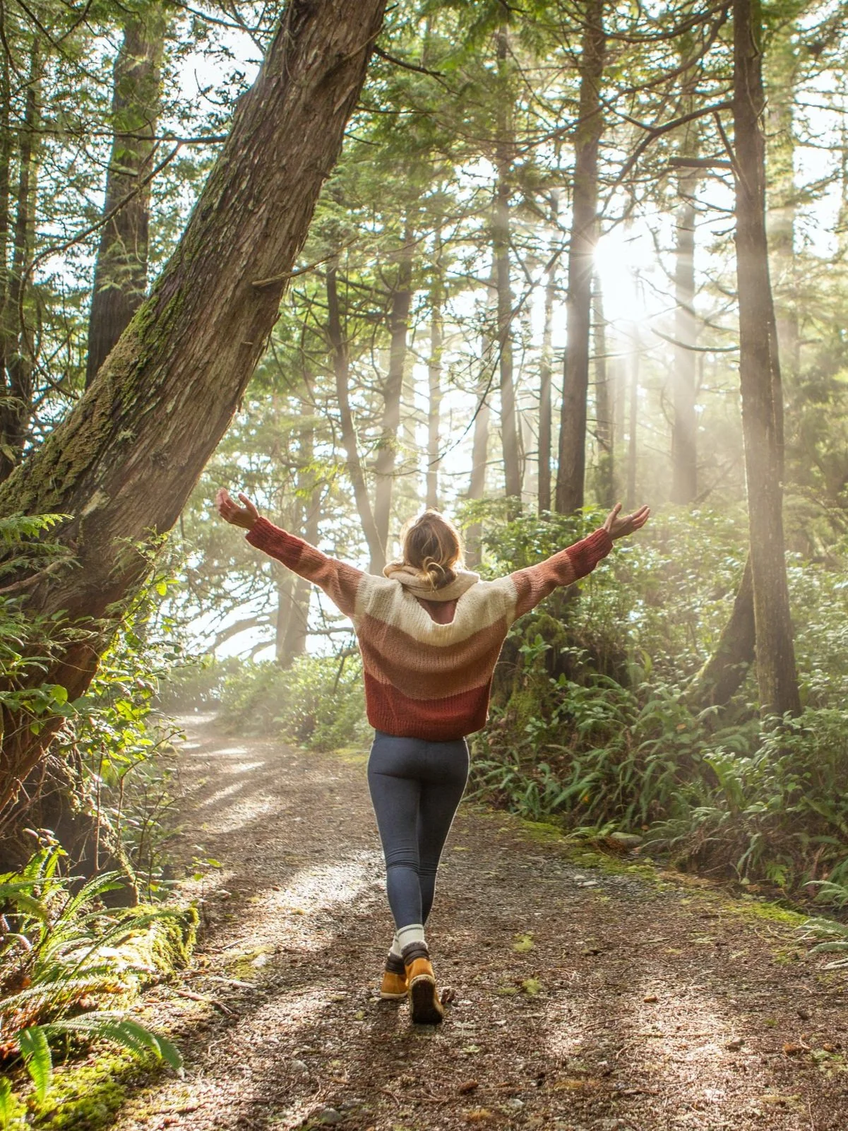 Eine Person geht mit ausgebreiteten Armen auf einem Waldweg, umgeben von hohen Bäumen, Sonnenlicht scheint durch das Geäst.
