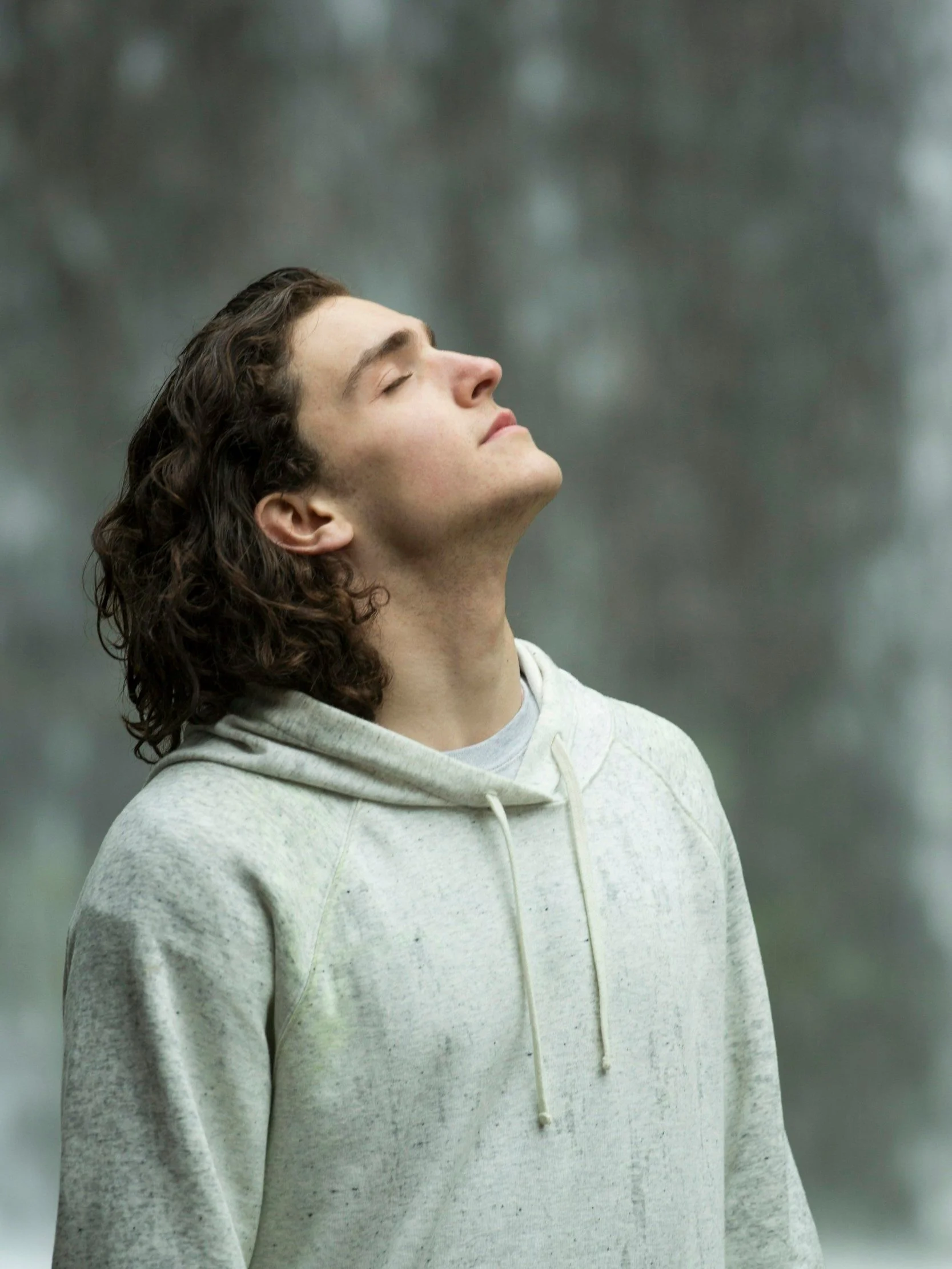 Ein junger Mann mit langen, lockigen Haaren trägt einen hellgrauen Hoodie und steht mit geschlossenem Blick in einer natürlichen Umgebung.