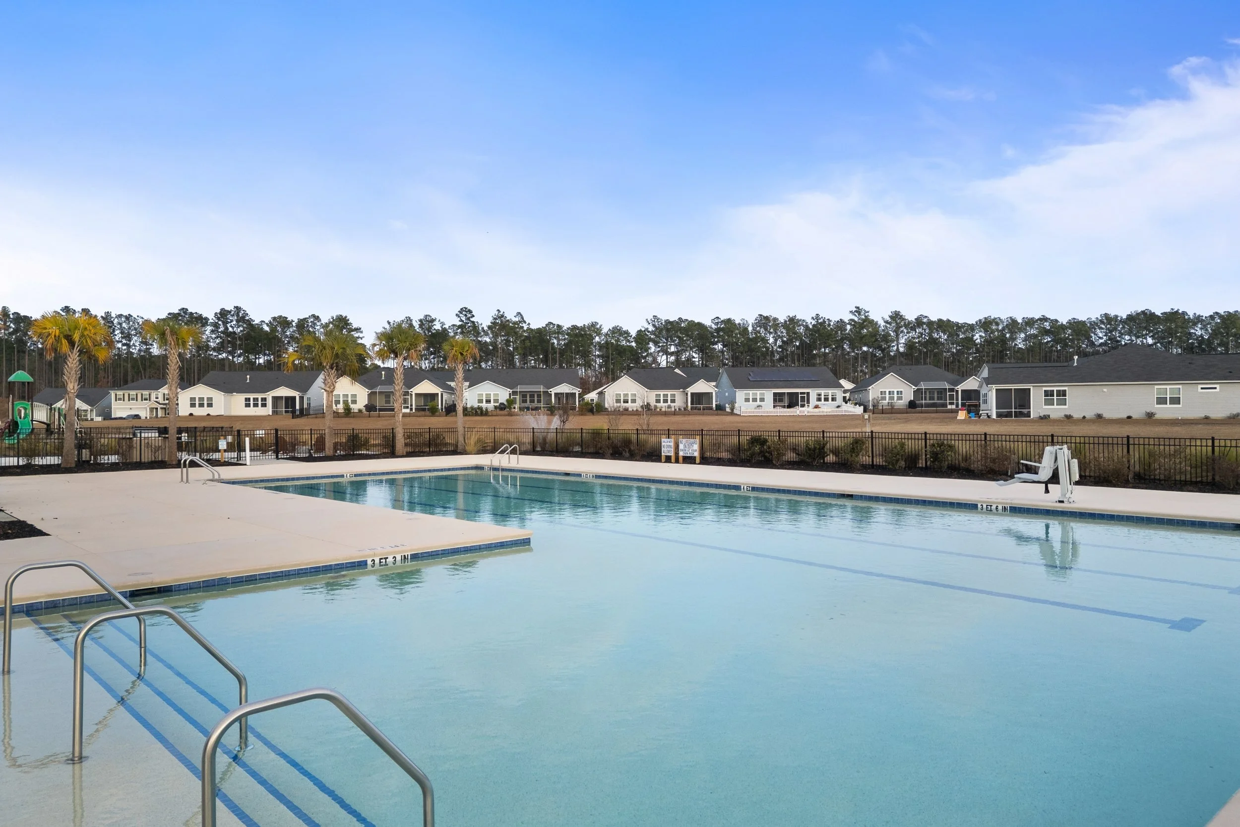 Empty outdoor community swimming pool with a few steps leading into the water, surrounded by a concrete deck, black metal fence, palm trees, and suburban houses in the background under a blue sky.