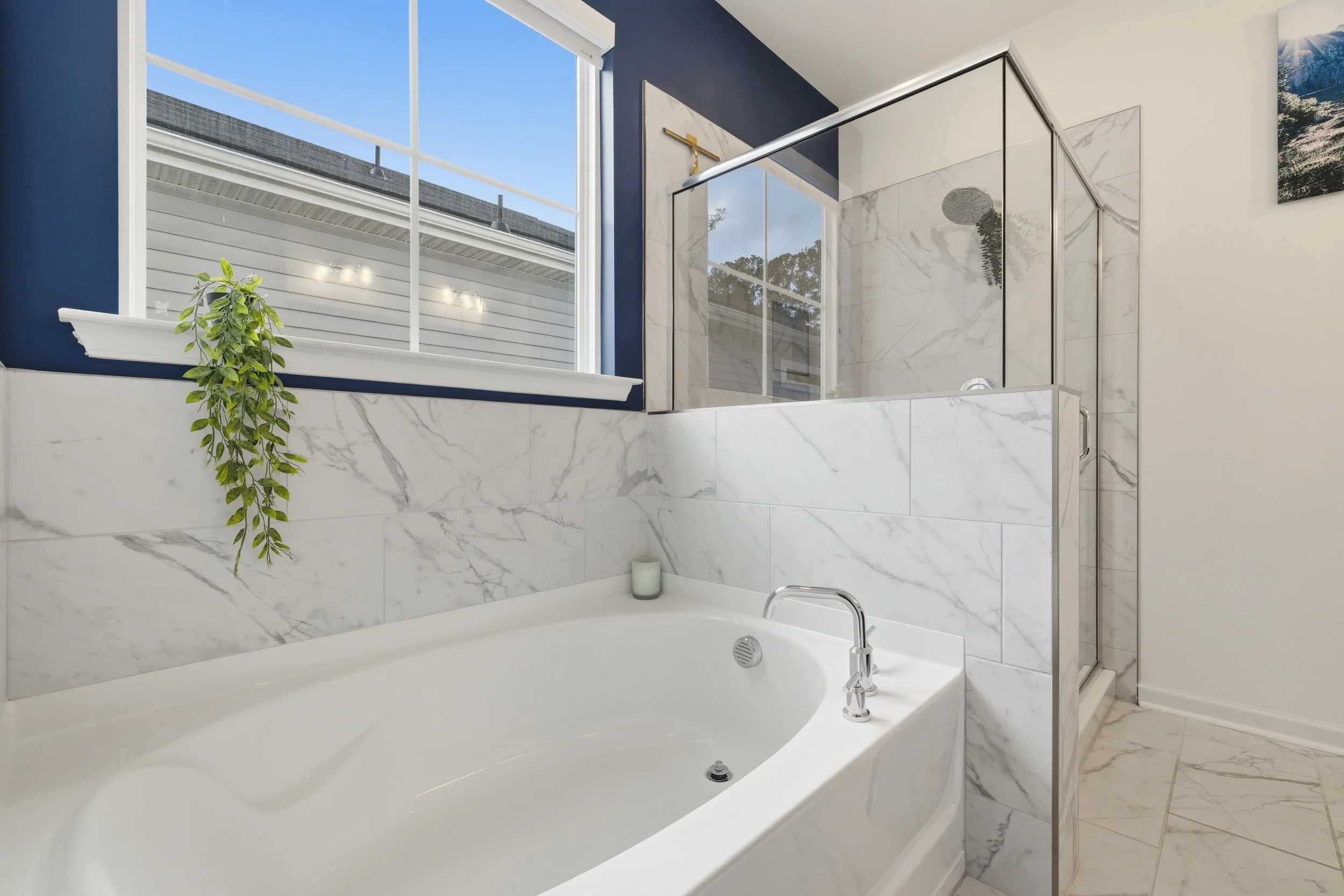Modern bathroom with a white bathtub, marble walls, a glass-enclosed shower, and a window with a plant on the windowsill.