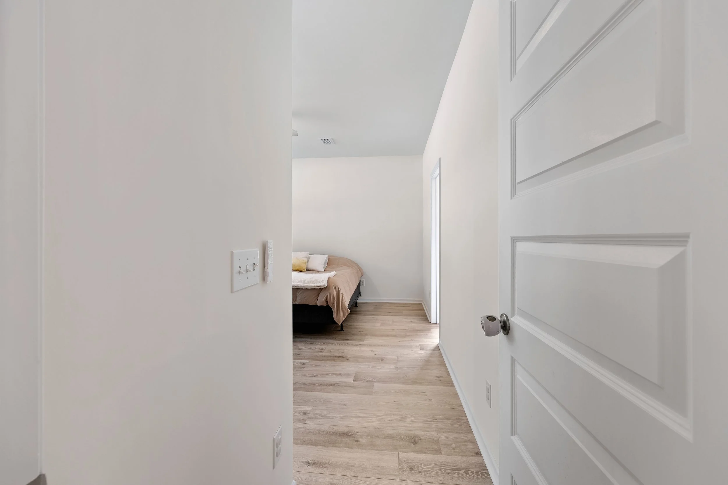 A view through a doorway into a room with a bed and beige bedding, a pillow, and a white wall in the background, with hardwood floors.