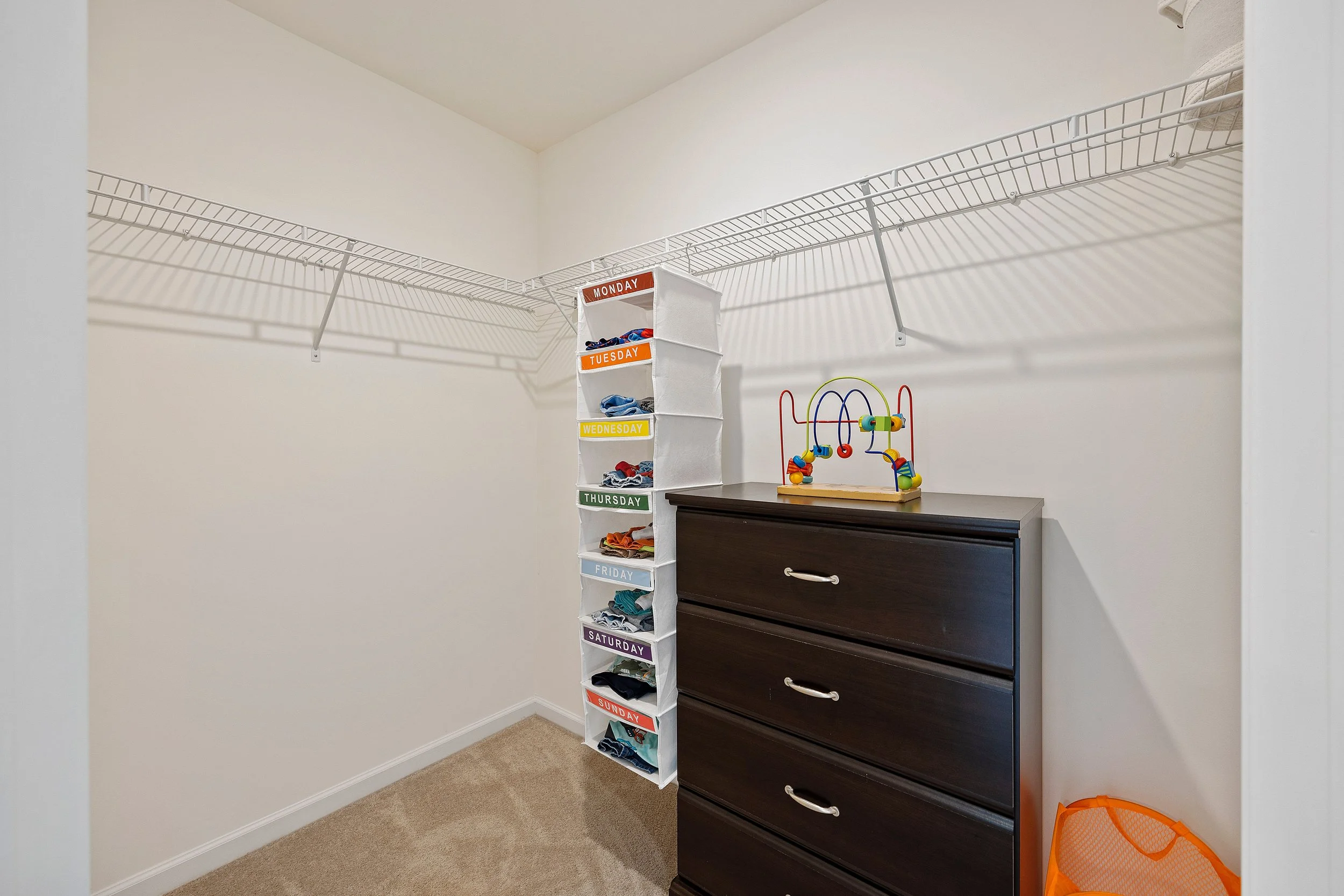 Empty walk-in closet with white wire shelving, colorful fabric storage bin with days of the week labels, a dark wooden dresser, and a small colorful bead toy on top.