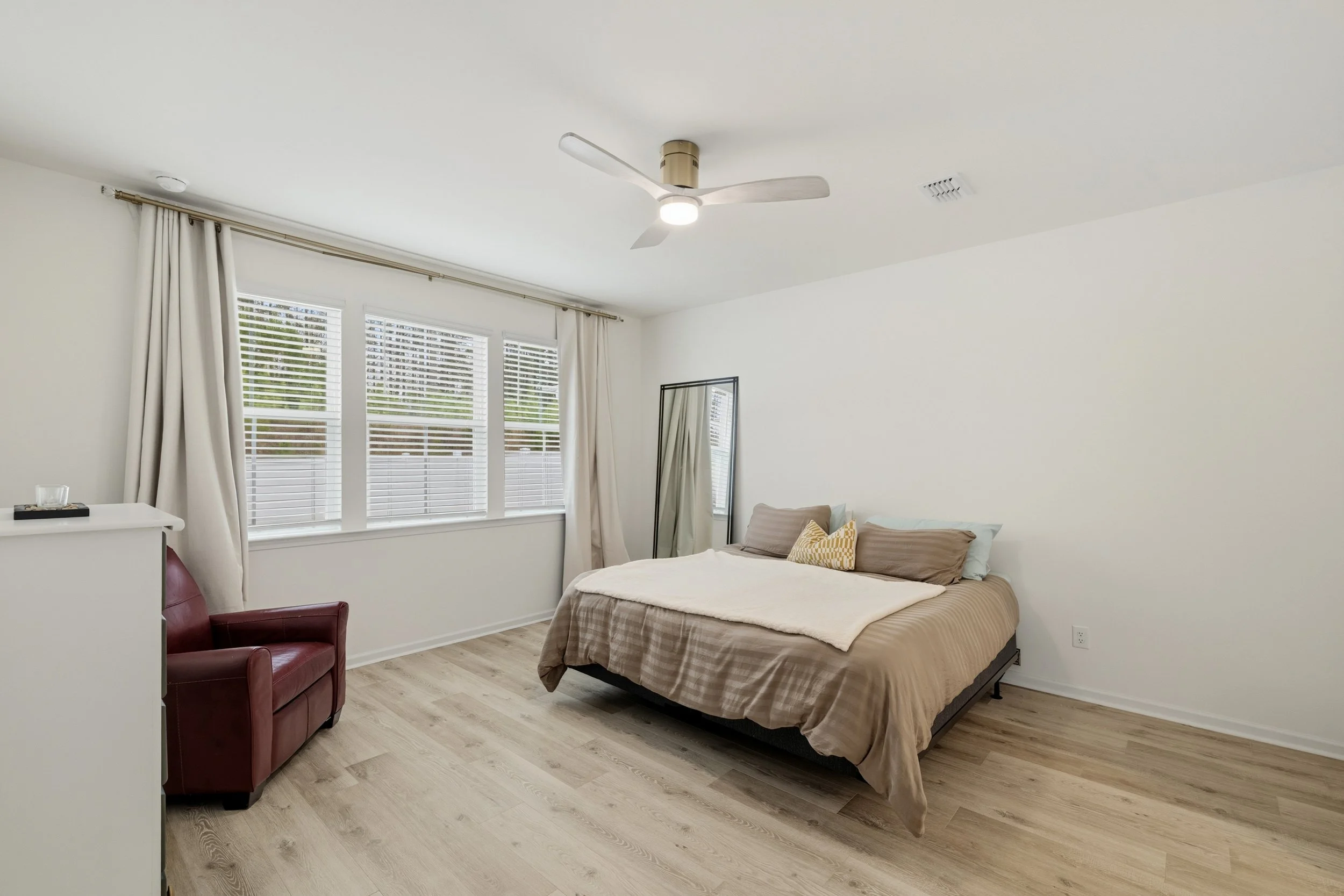 Minimalist bedroom with a bed, pillows, mirror, red armchair, and large window with white blinds and curtains.