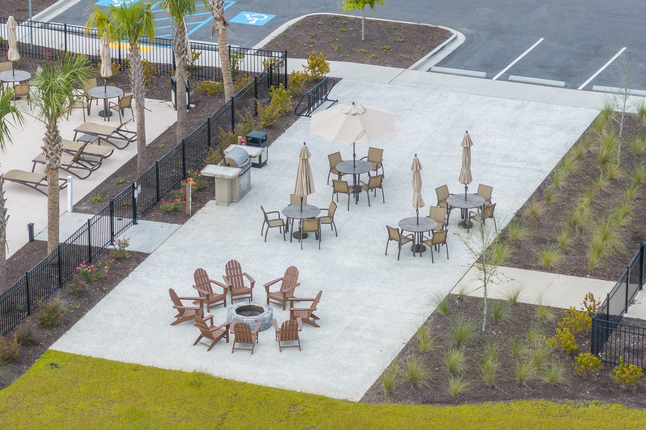 An outdoor patio area with several tables, chairs, and umbrellas, surrounded by landscaped garden beds and a parking lot in the background.