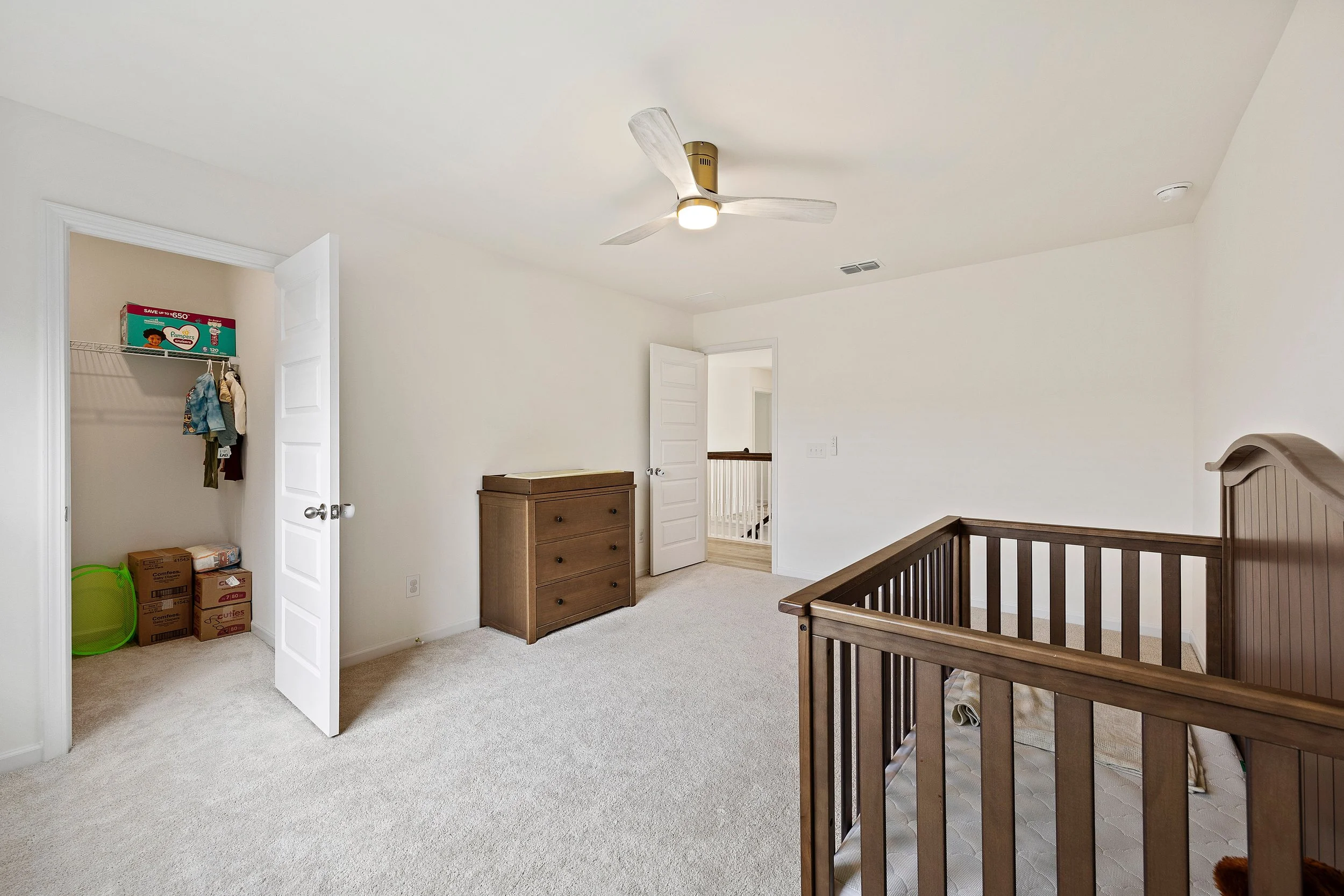 Empty nursery room with a wooden crib, dresser, ceiling fan, doorway, and an open closet with boxes and clothing.