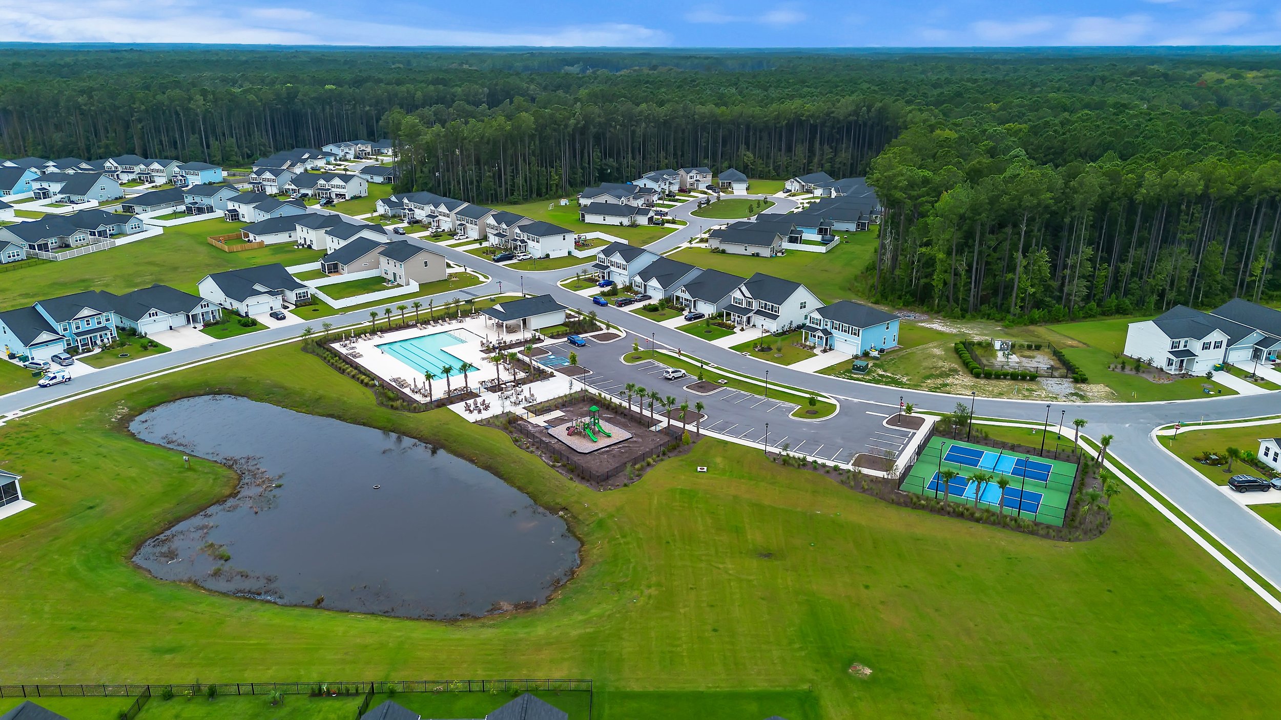 Aerial view of a suburban neighborhood featuring houses, a community pool, a playground with slides, a tennis court, a pond, and roads connecting the housing area, with a wooded area in the background.