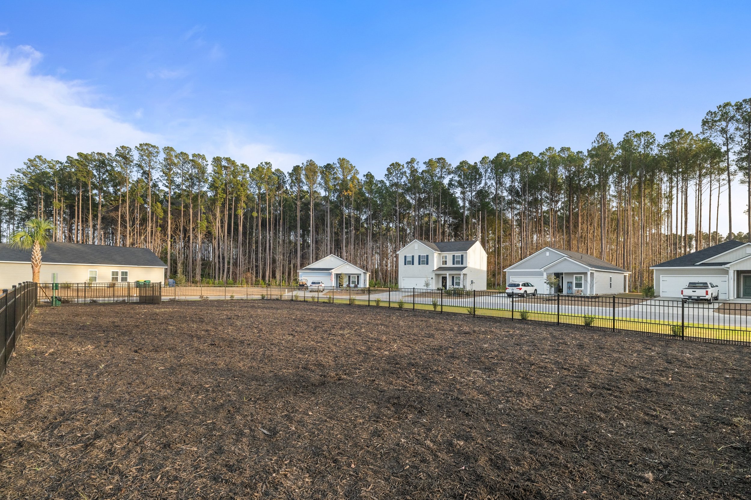 A fenced, cleared piece of land with several modern houses and a row of tall trees in the background under a clear blue sky.