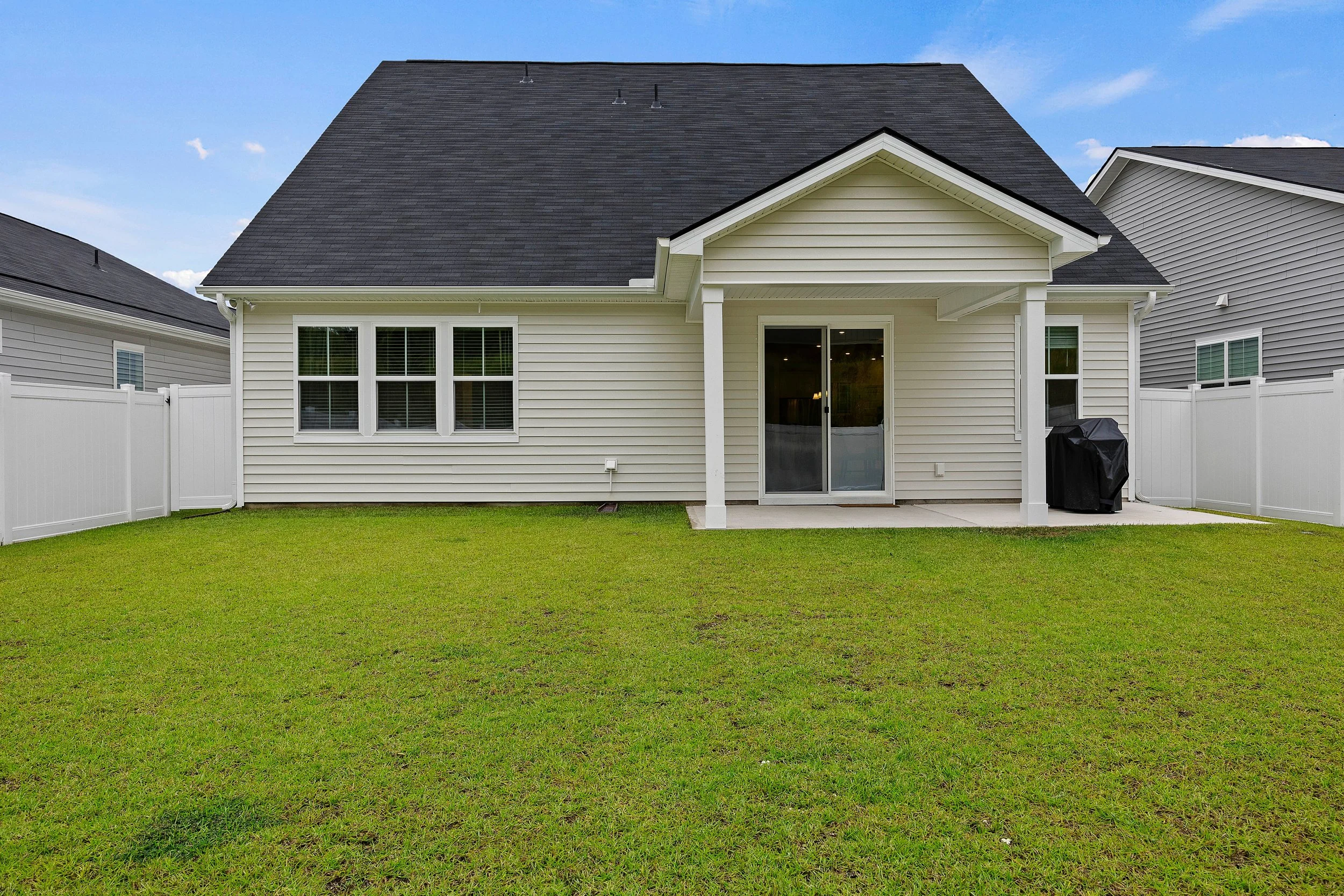 Backyard with a white house, black roof, sliding glass door, two windows, green grass, and a white privacy fence.