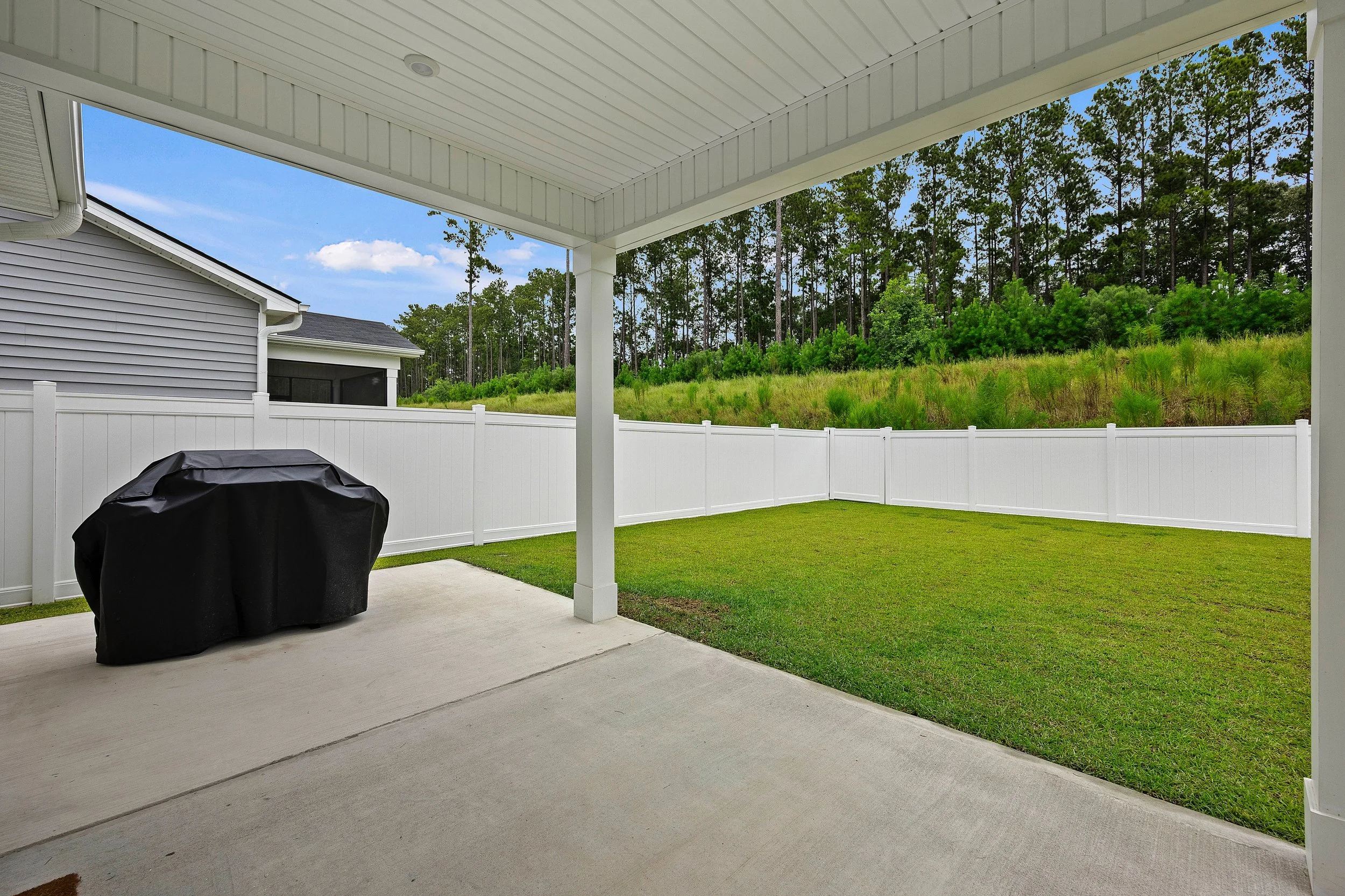 Backyard with patio, barbecue grill covered with a black cover, white fence, green grass, and trees on a hill under a blue sky with clouds.
