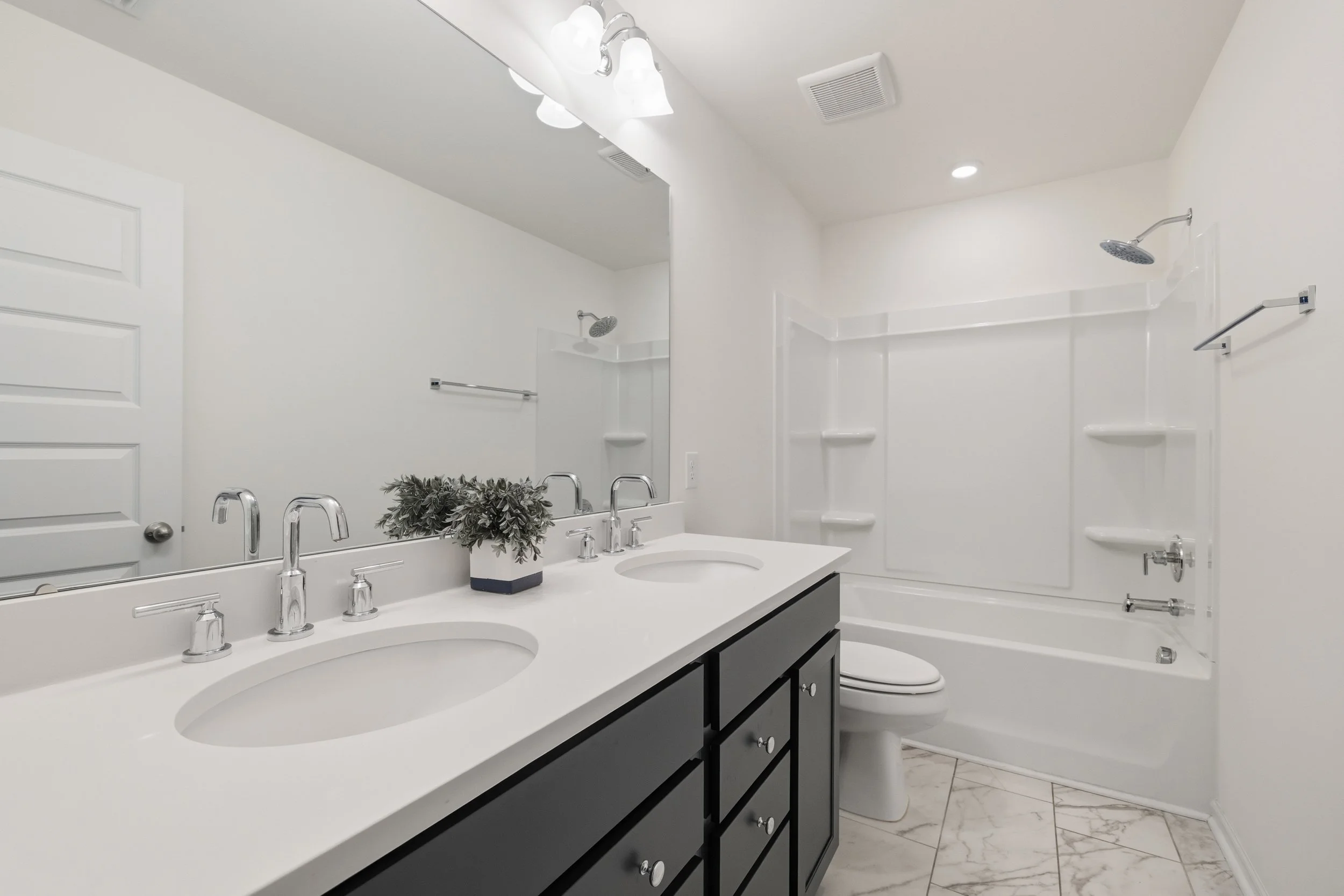 Clean bathroom with dual sinks, black vanity, large mirror, white bathtub and shower with built-in shelves, white walls, marble floor, and ceiling light fixtures.