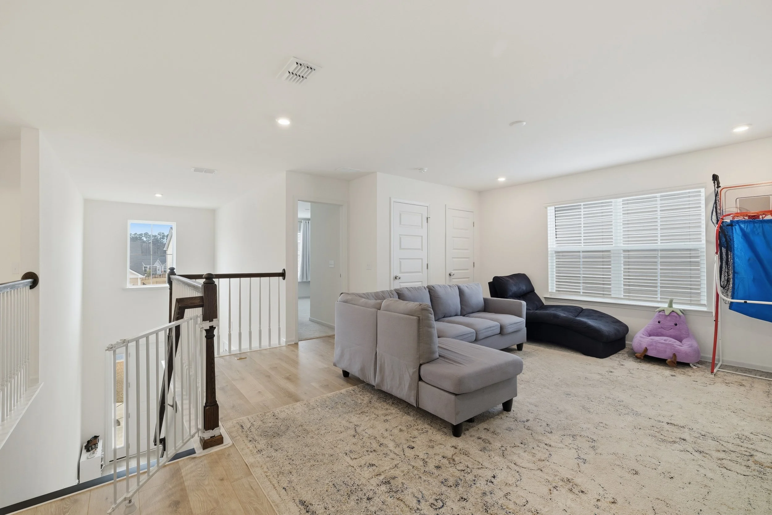 A bright living room with a gray sectional sofa, a black chaise lounge, a large window with blinds, a plush pink bean bag, and a blue and orange gym set in the corner.