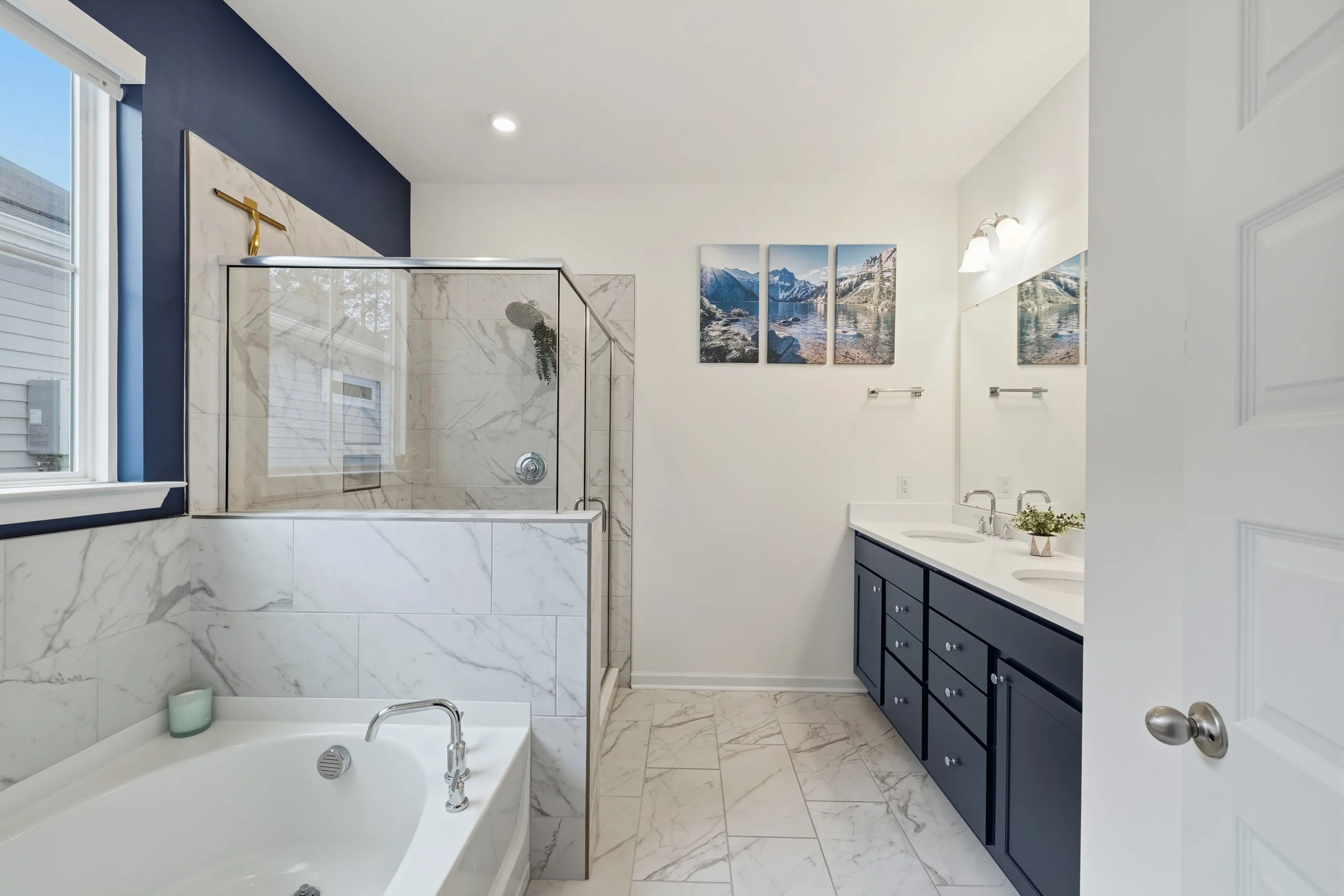 Bathroom with a marble bathtub and shower, navy blue vanity with double sinks, three-panel mountain landscape art, and a window with blue walls.