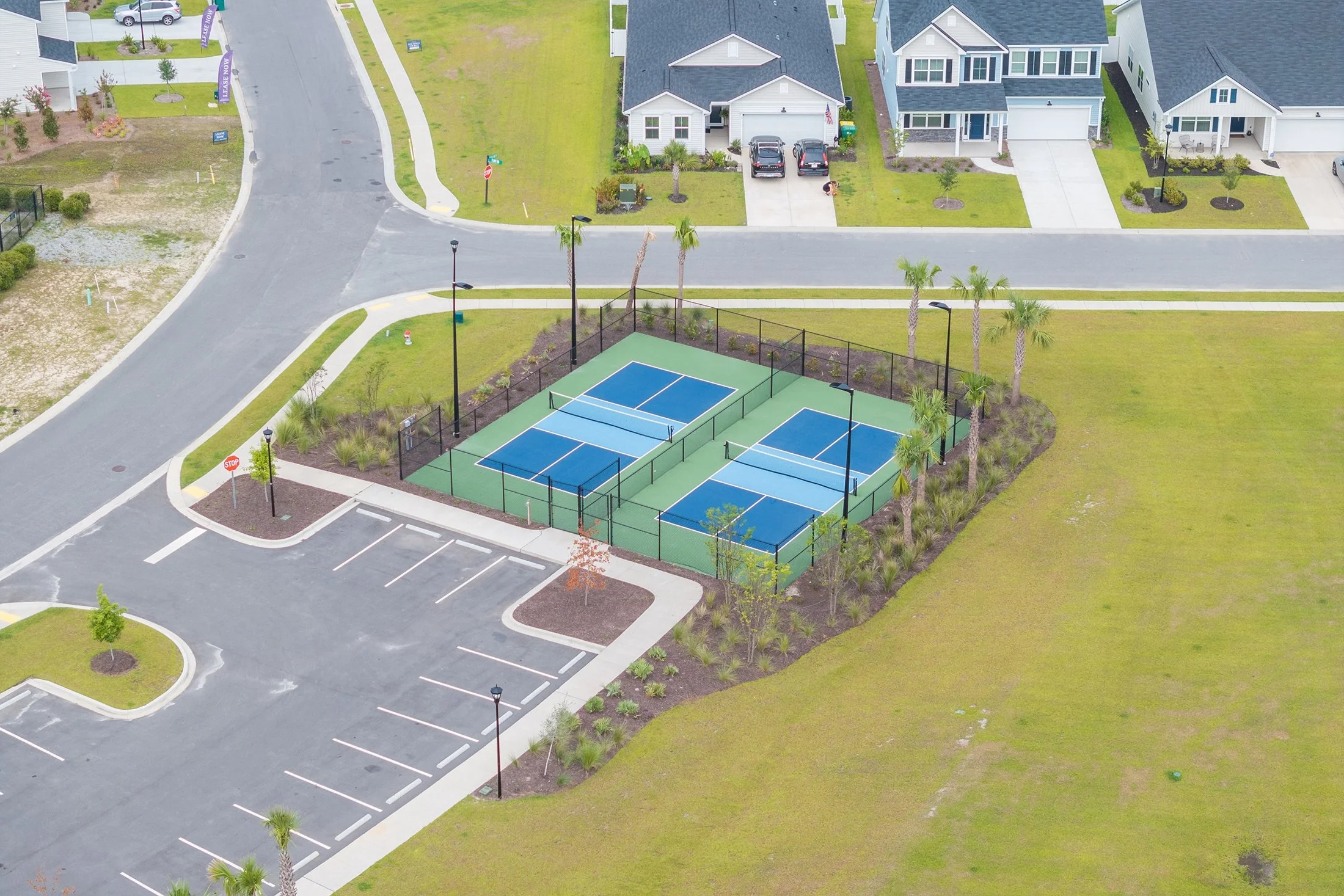 A community outdoor tennis court area with four courts, surrounded by palm trees and landscaped bushes, adjacent to parking spaces and residential houses in the background.