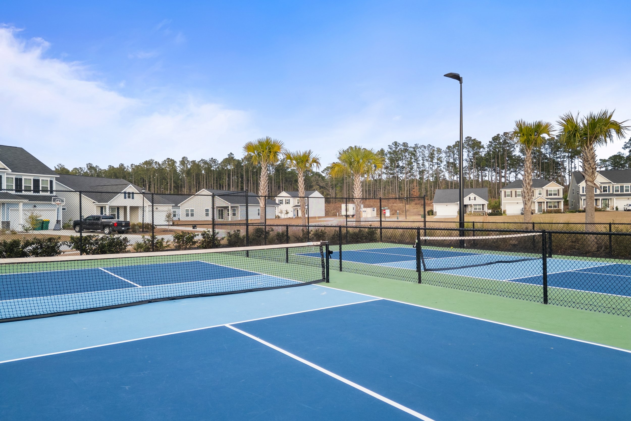 Empty outdoor tennis courts with blue and green surfaces, surrounded by a black chain-link fence, with trees and residential houses in the background under a partly cloudy sky.