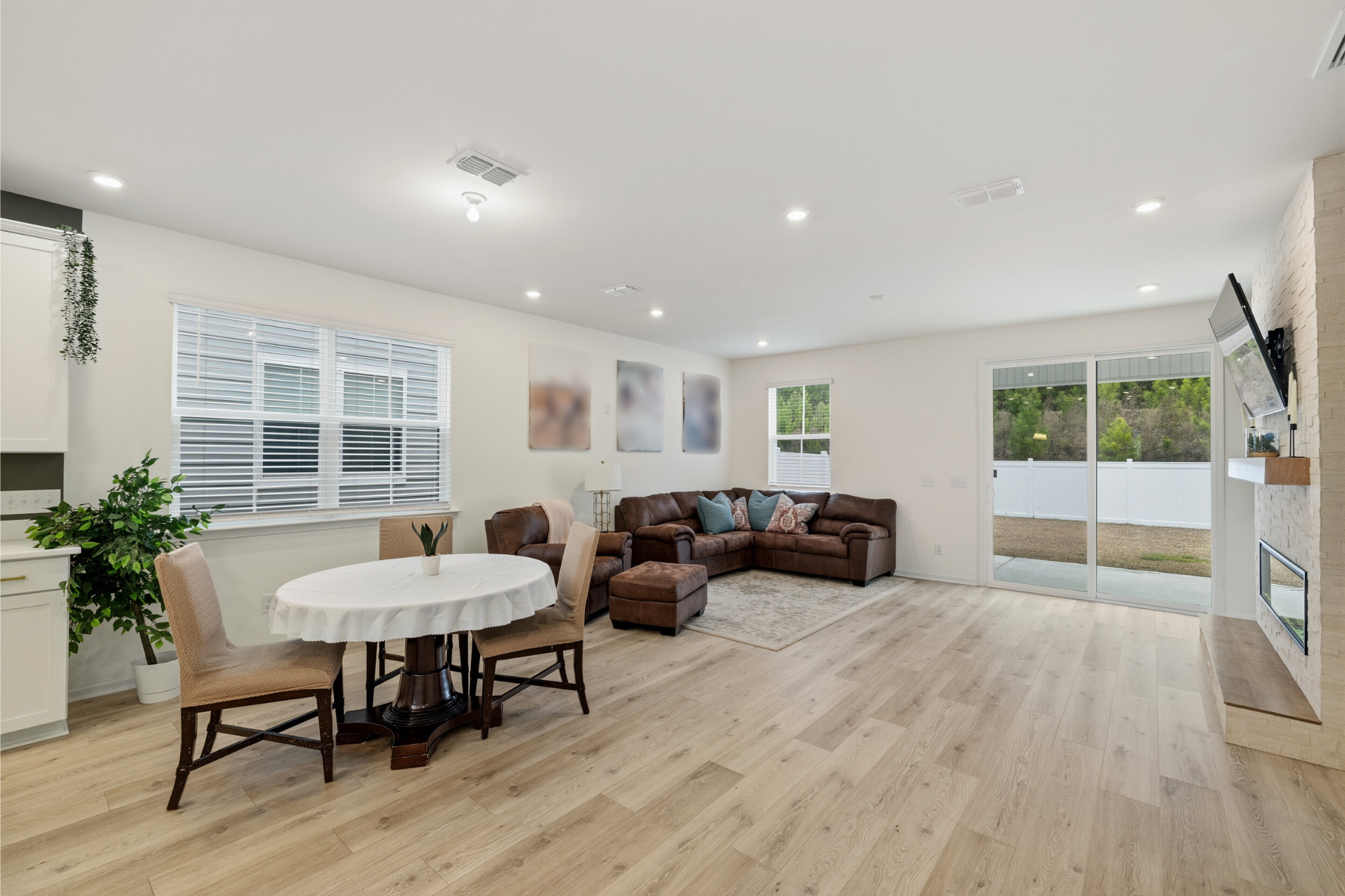 Open living room with hardwood floors, a brown sectional sofa with cushions, a sliding glass door leading outside, a wall-mounted TV, and a dining area with a round table and beige chairs, illuminated by ceiling lights.