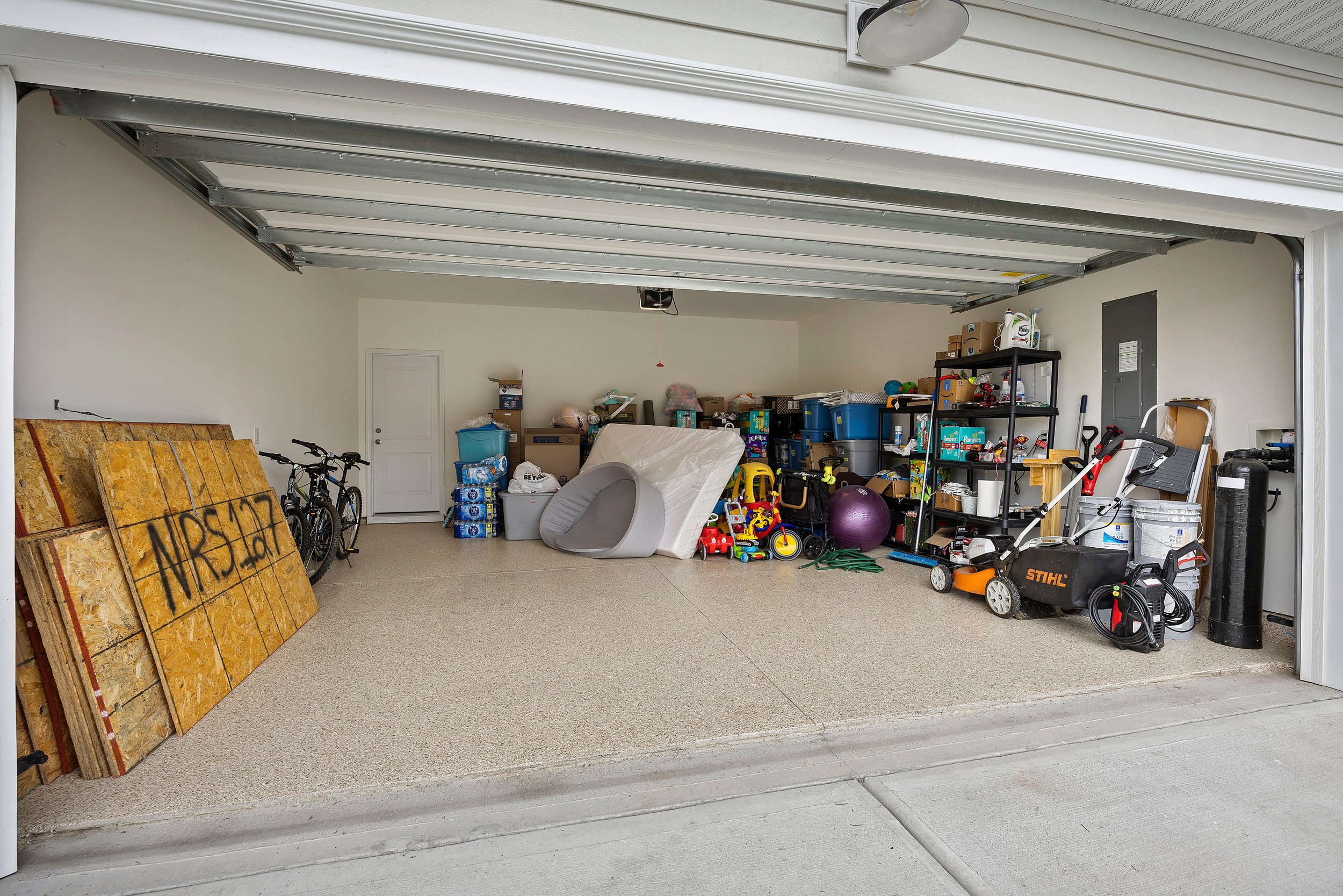 Inside a garage filled with storage bins, a lawnmower, toys, a purple exercise ball, bikes, and construction materials.