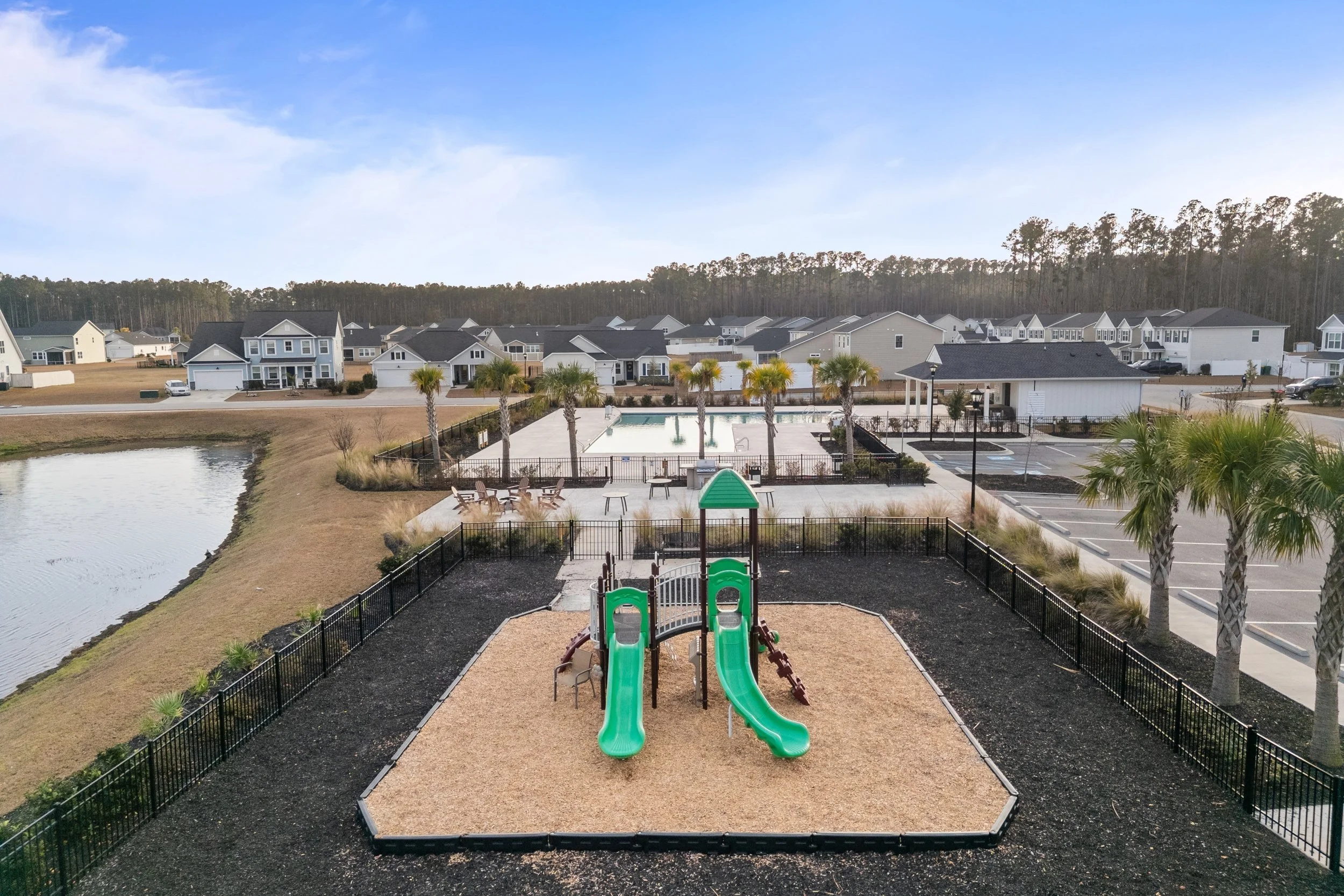 A playground with a green slide and climbing structure surrounded by black fencing, with a row of palm trees behind it. In the background, there is a swimming pool area with lounge chairs, a seating area, and parking lot, within a residential communi