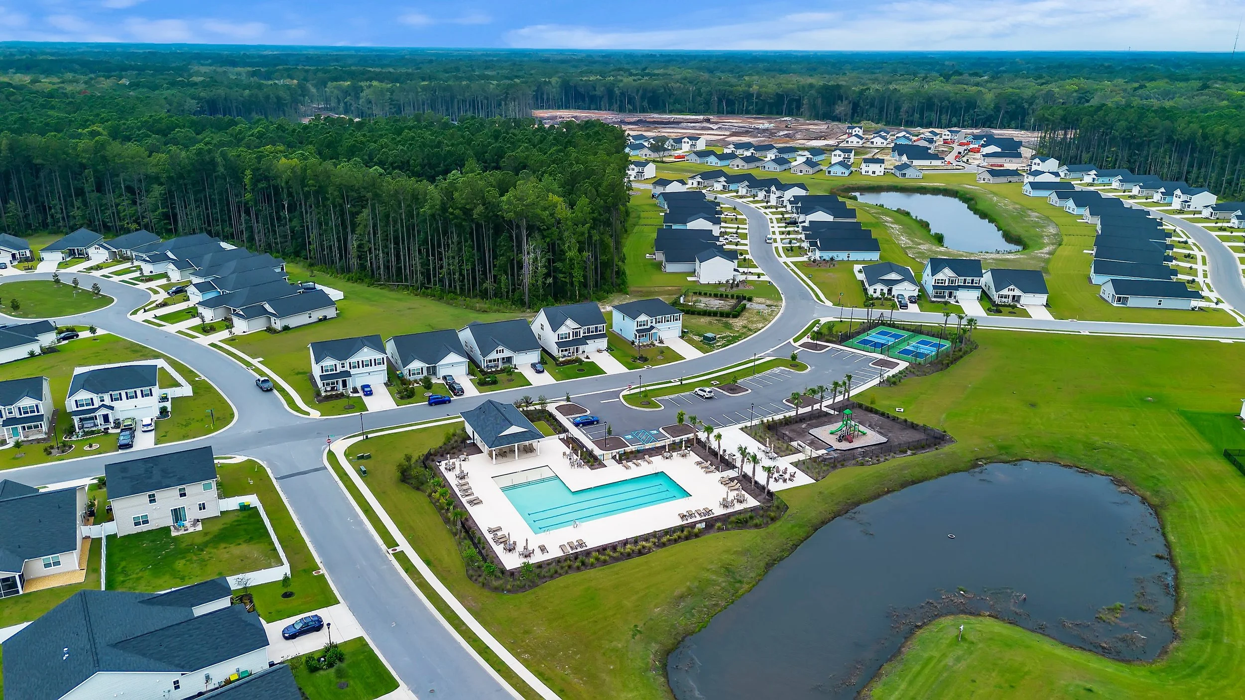 Aerial view of a residential community with houses, a swimming pool, playground, tennis courts, pond, and green lawns, surrounded by forest.