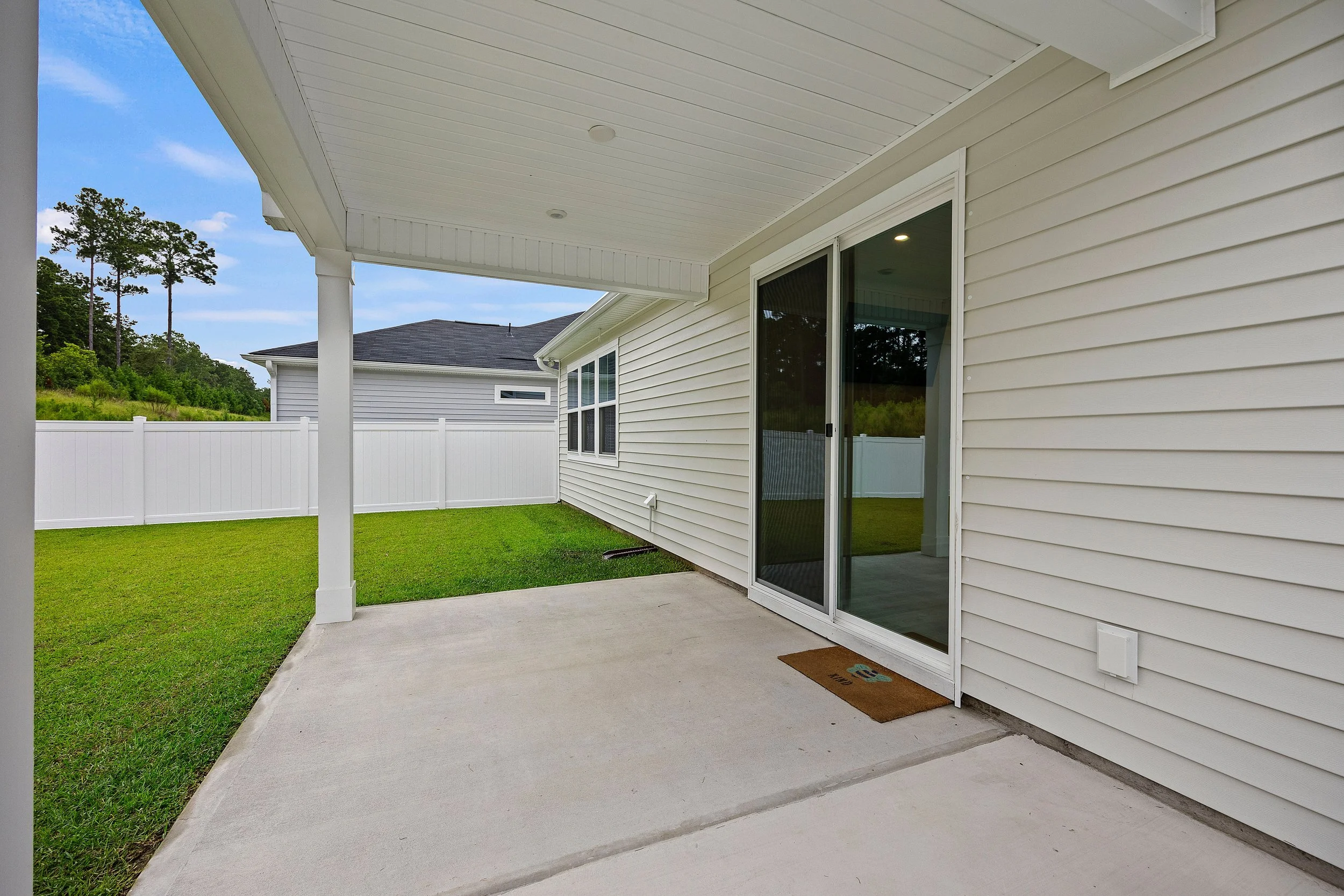 Backyard patio area with concrete floor, white vinyl siding, sliding glass door, lawn, white vinyl fence, and a neighbor's house with trees in background.