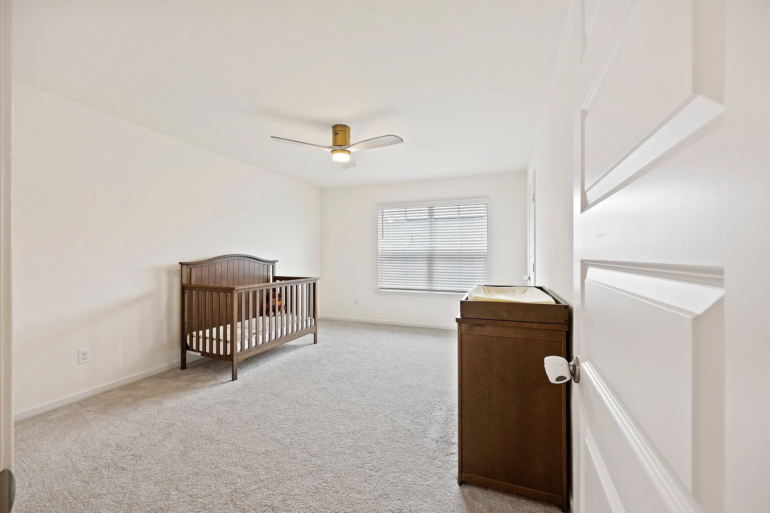 Empty baby nursery with crib, changing table, window with blinds, ceiling fan, and beige carpeting.