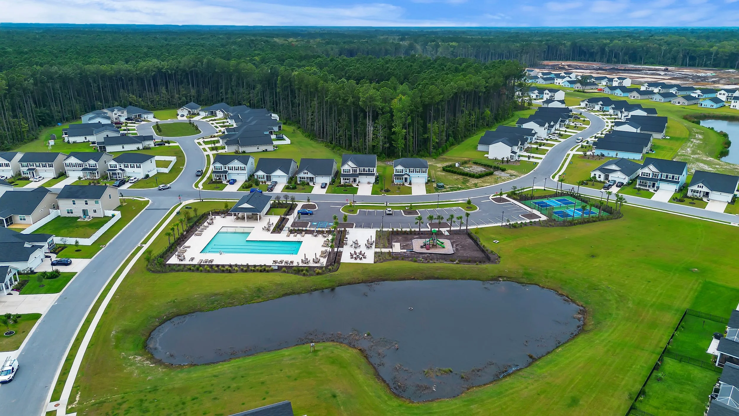 Aerial view of a residential community with houses, a swimming pool, playground, tennis courts, and a pond near green forest.