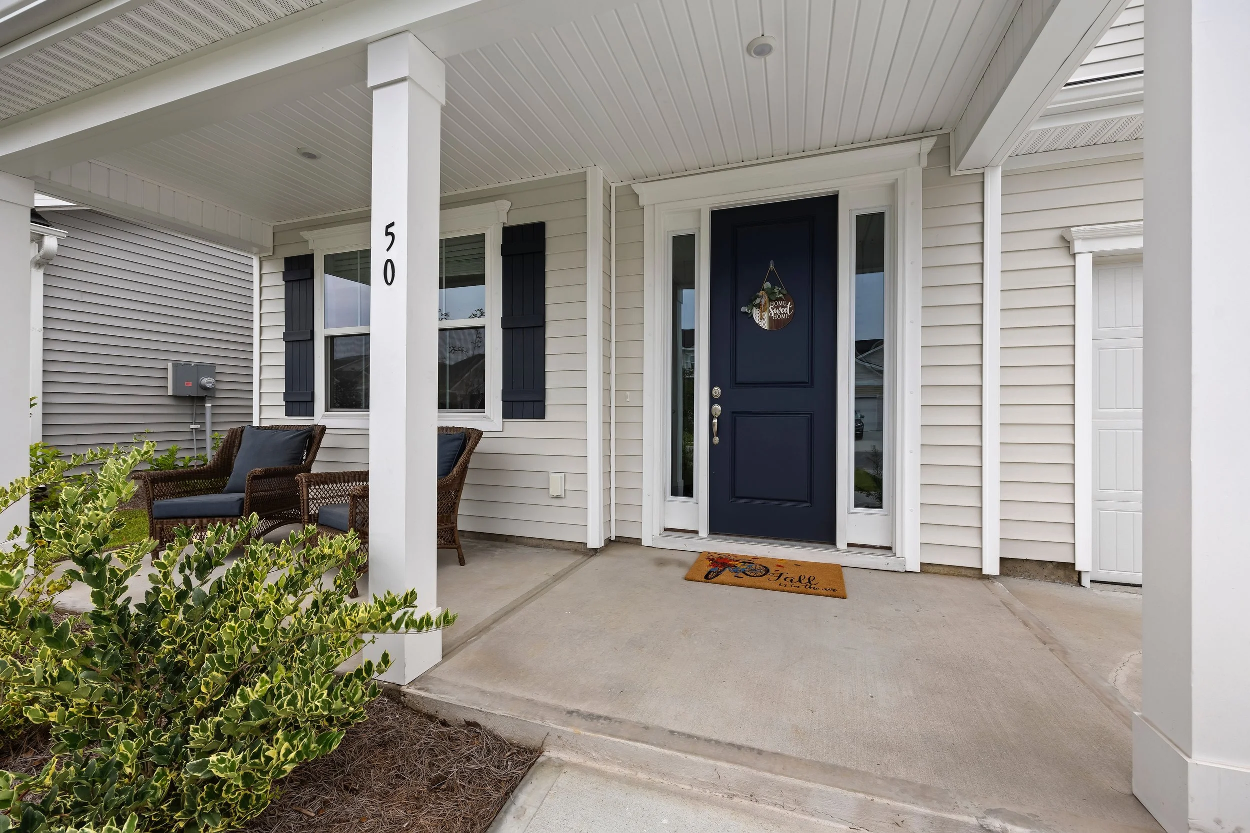 House entrance with blue door, white siding, black shutters, and porch with chairs and a welcome mat.
