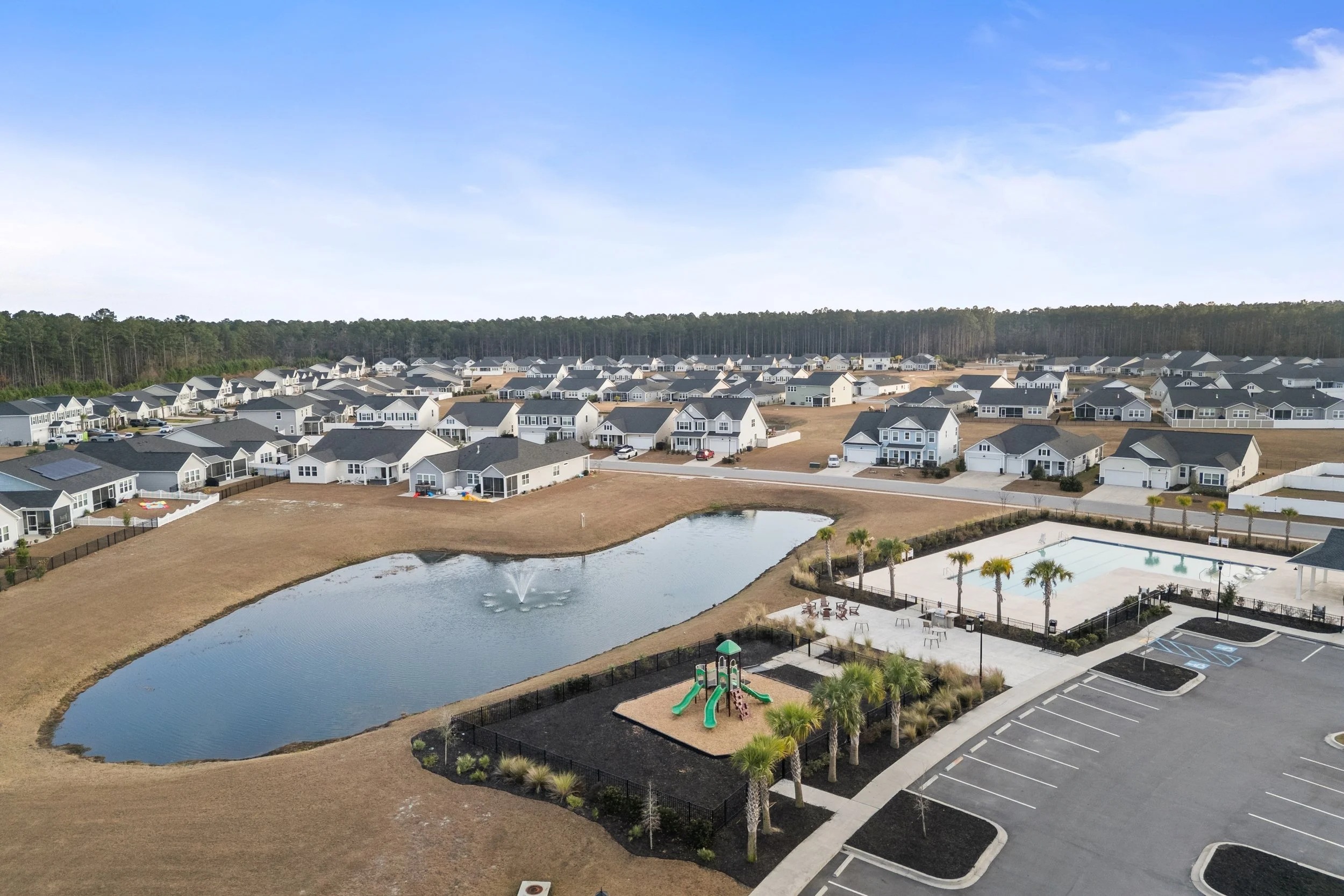 A residential neighborhood with multiple houses, a pond with a fountain, a playground with slides, a tennis court, and a parking lot, all surrounded by trees under a partly cloudy sky.