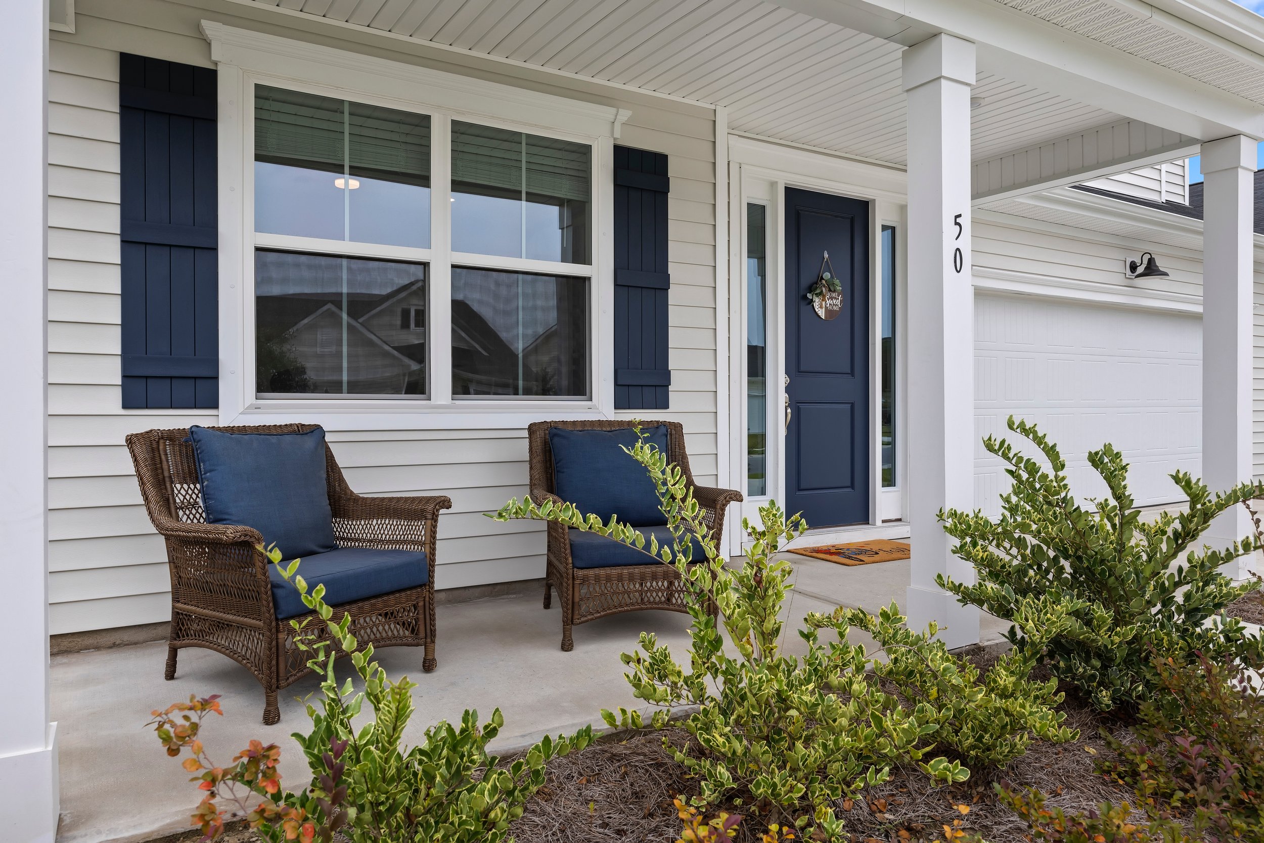 Front porch of a house with two wicker chairs with dark blue cushions, a dark blue front door with a decorative wreath, and a window with navy shutters, surrounded by greenery.