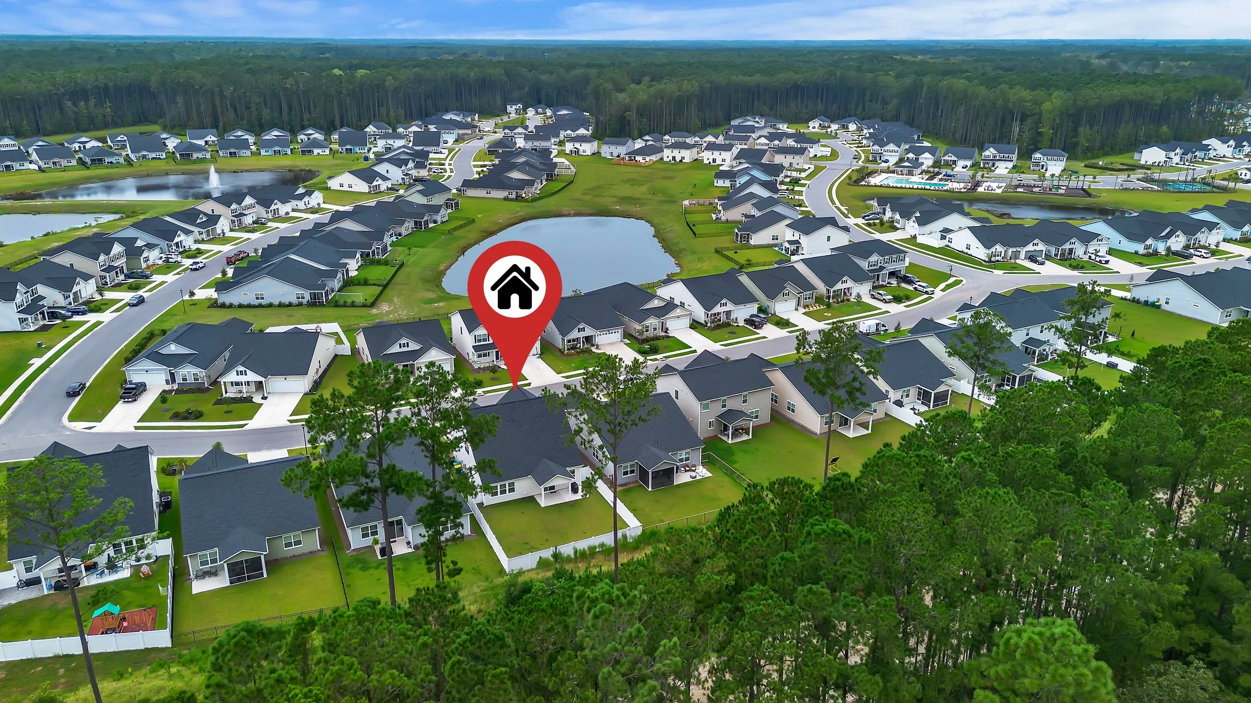Aerial view of a suburban neighborhood with multiple houses, some with fenced yards, a pond, and green trees, with a red marker indicating one specific house.
