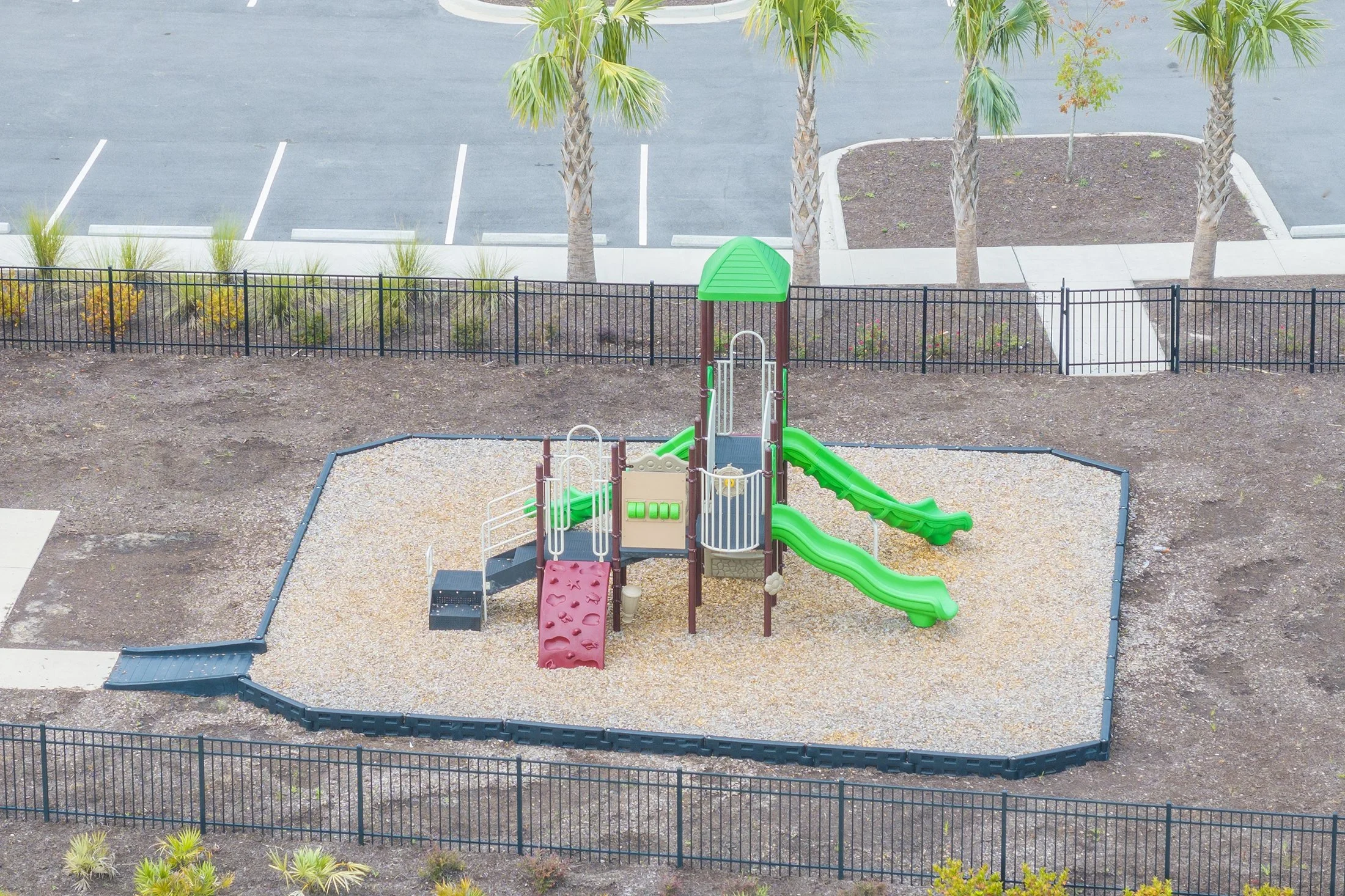Empty playground with green slides, climbing wall, and green roof, enclosed by a black fence. Surrounding area has trees and parking lot.