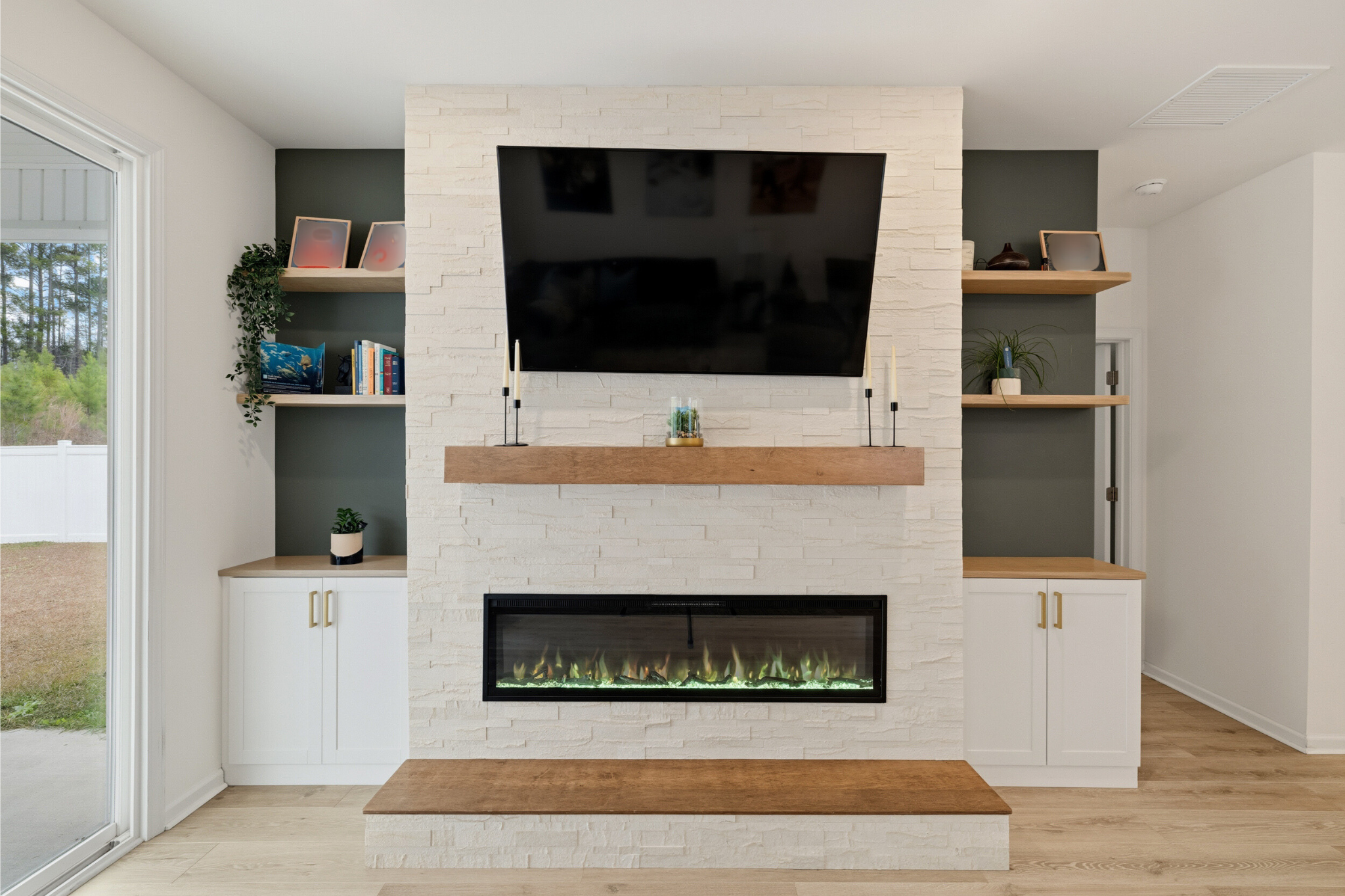 Modern living room with white brick fireplace, mounted flat-screen TV, wooden shelves, and built-in cabinets, with sliding glass door to the left showing outdoor greenery.