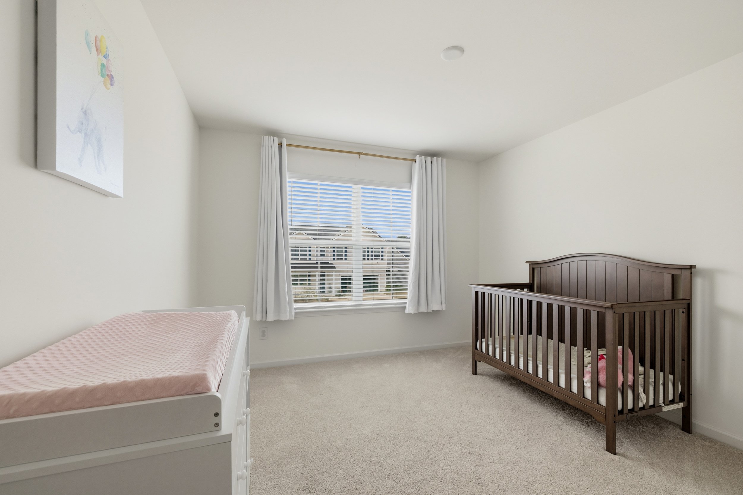 A nursery with white walls, a window with white curtains, a white changing table, and a dark brown crib with a pink stuffed animal inside.
