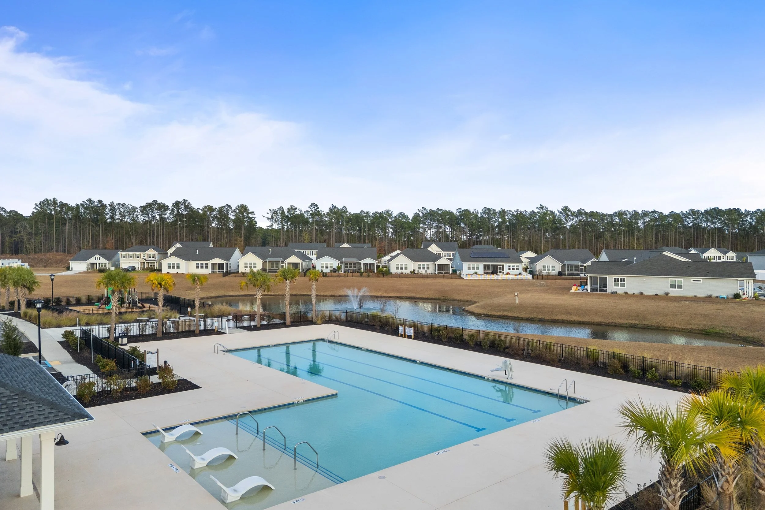 Community swimming pool with lounge chairs and a pond in the background, surrounded by residential houses and palm trees under a blue sky.
