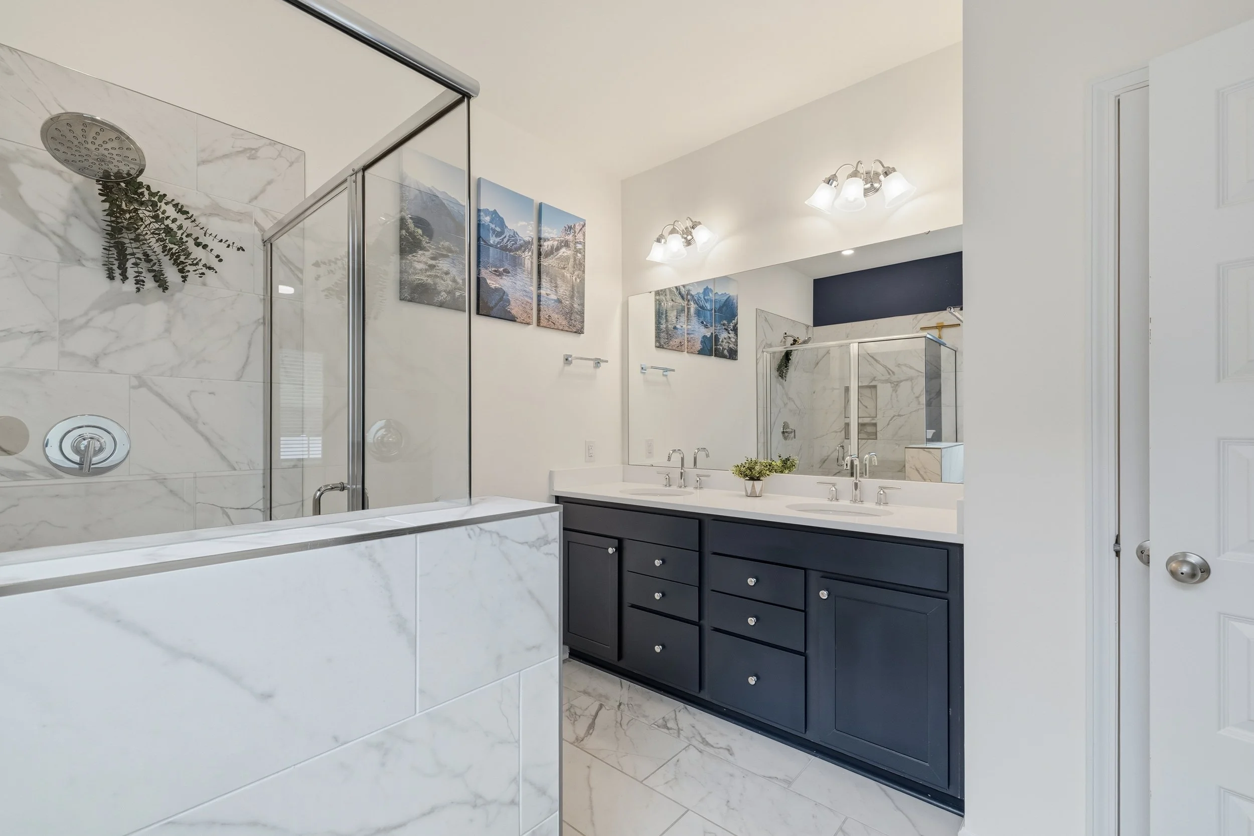 Modern bathroom with marble shower, dark blue vanity, large mirror, white countertops, and mountain landscape artwork