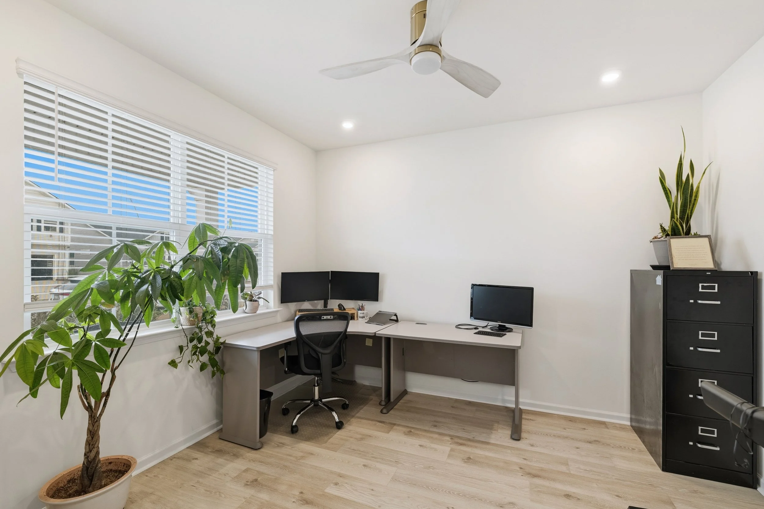 Clean modern home office with a large window, a black filing cabinet, a desk with dual monitors and a desktop computer, a black swivel chair, potted plants, and hardwood flooring.