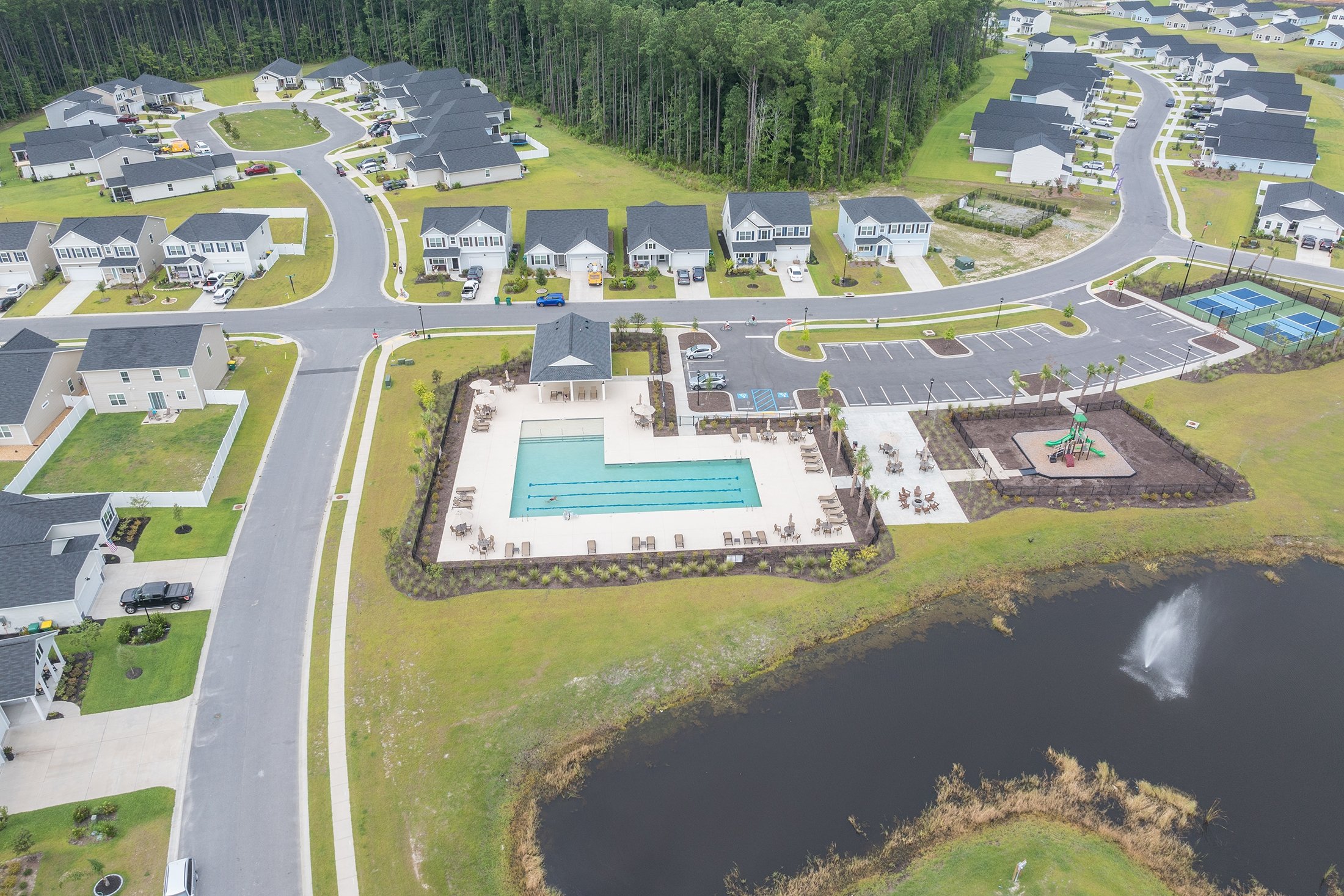 Aerial view of a residential neighborhood featuring a community swimming pool, playground, parking lot, tennis courts, and surrounding houses with well-maintained lawns and a pond with a fountain.
