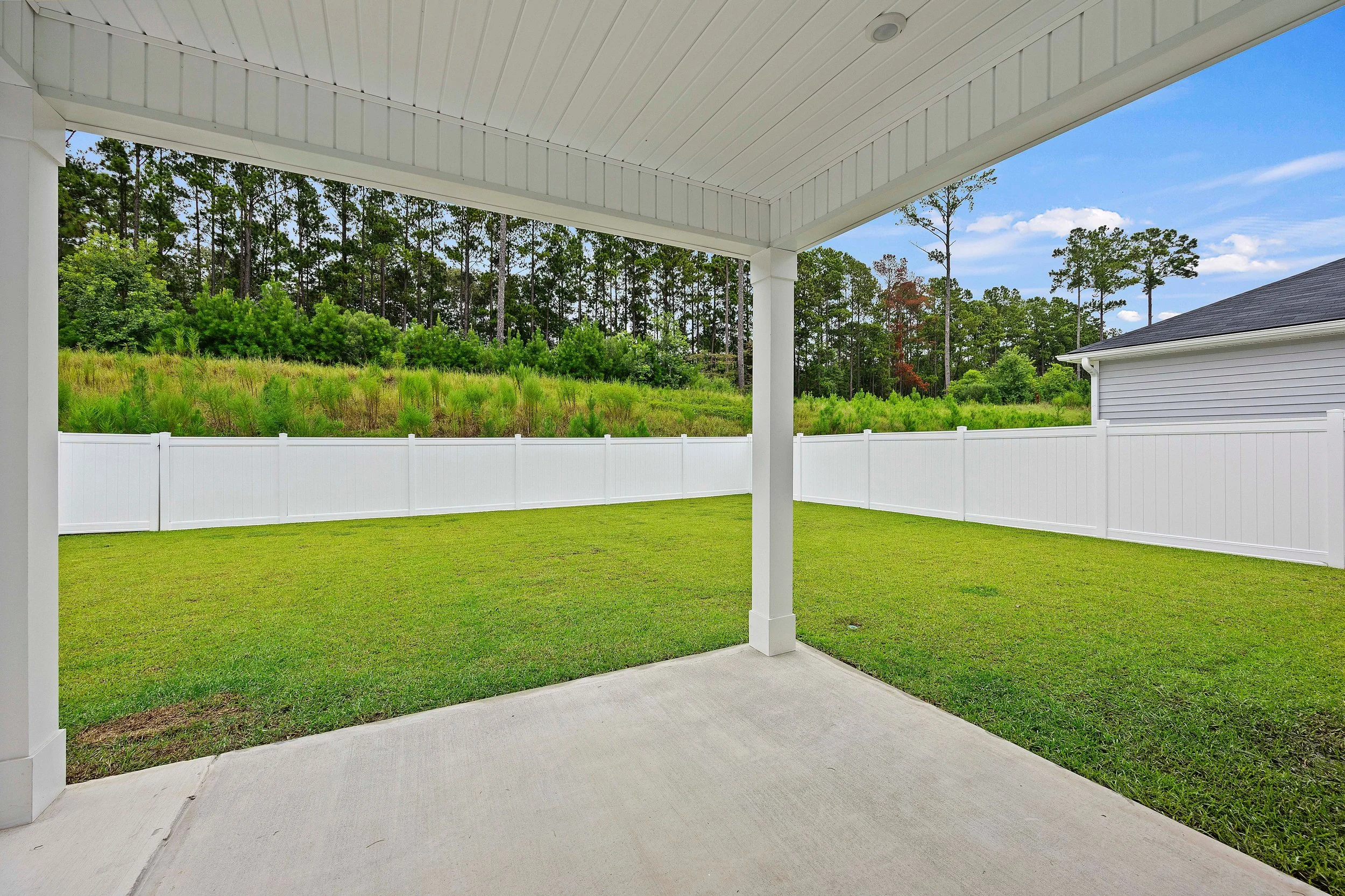 View from a covered patio looking out onto a grassy backyard enclosed by a white privacy fence, with trees and a partly cloudy sky in the background.