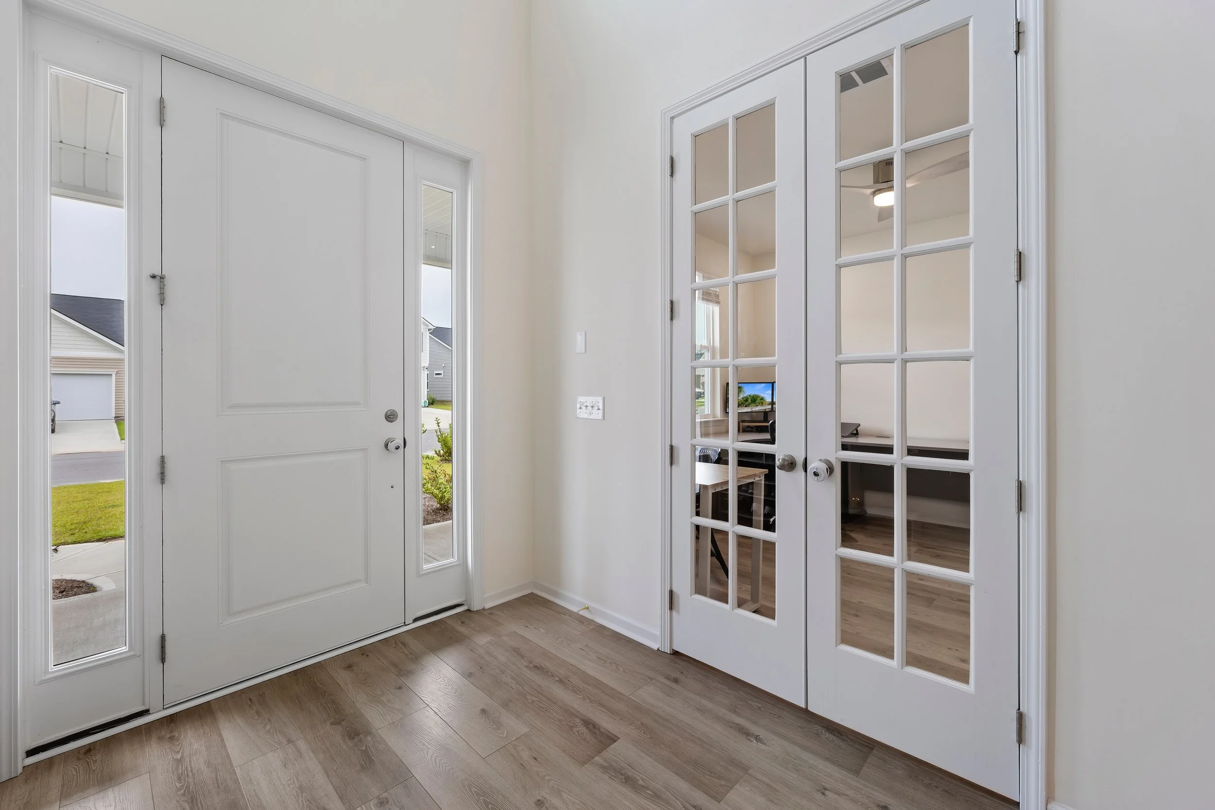 Entryway with white front door and glass-paneled French doors leading to another room, light-colored wood flooring, and partial view of outside through side windows.