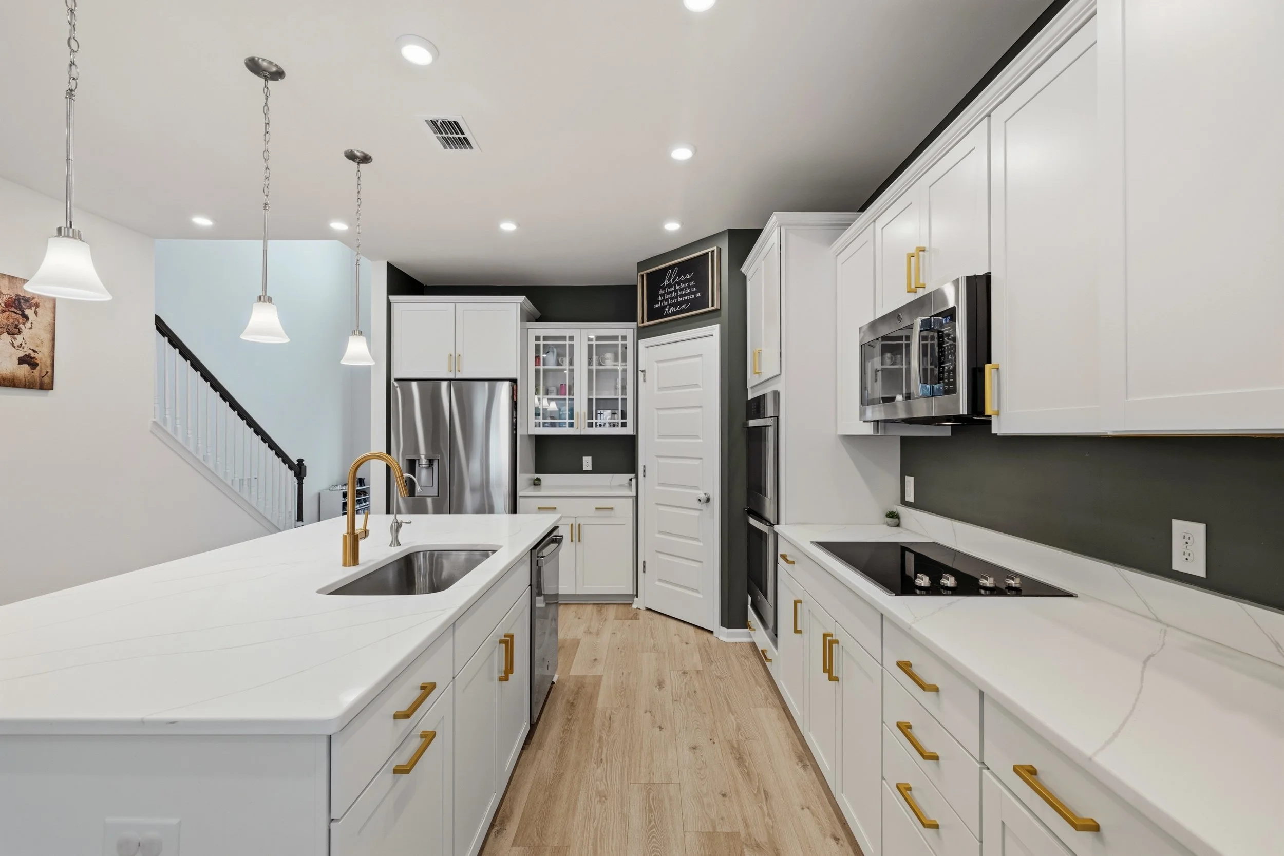 Modern kitchen with white cabinets, gold handles, stainless steel appliances, a white island with a gold faucet, and pendant lighting. There is a black accent wall and hardwood floors.