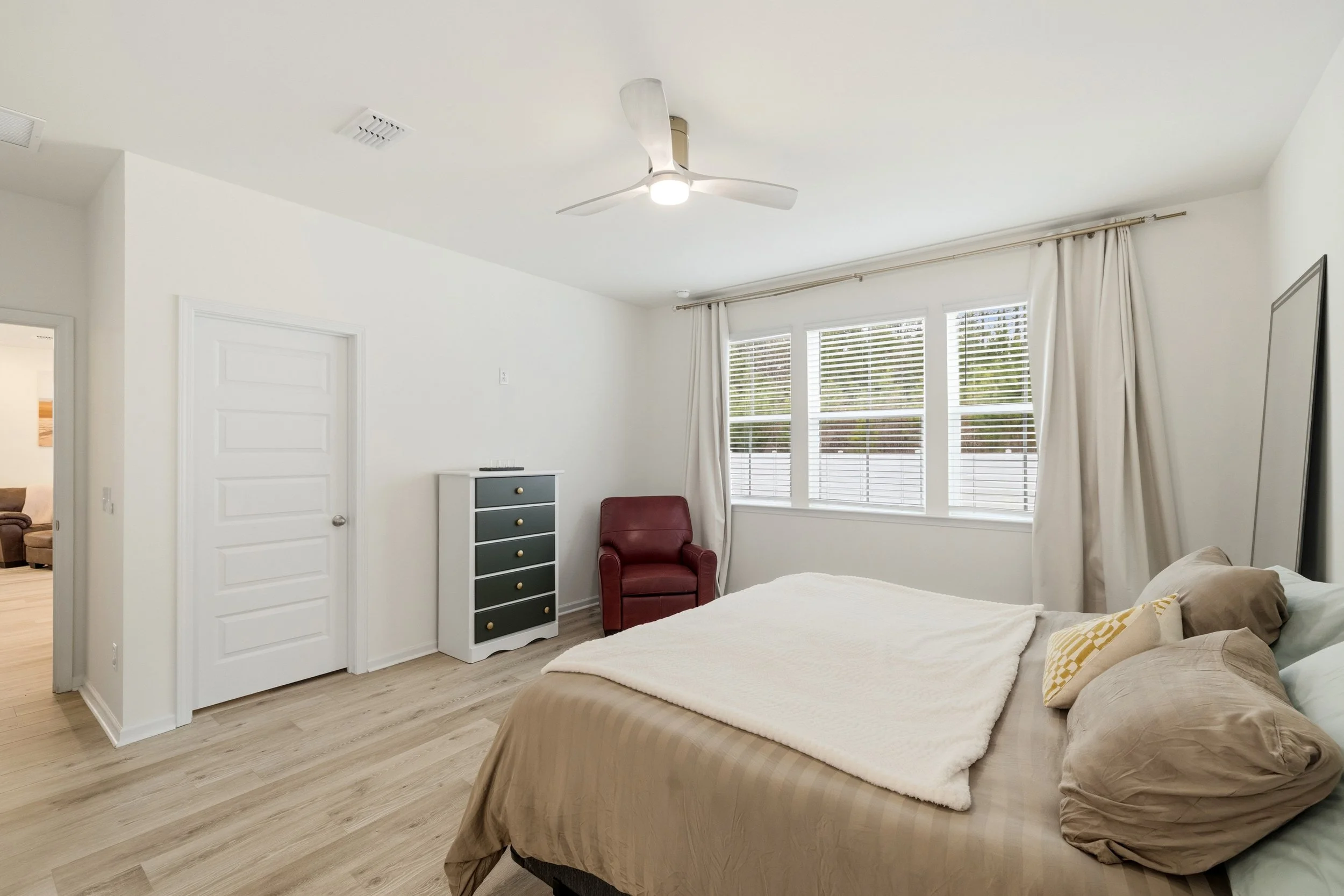 Bright bedroom with a bed, pillows, and a white blanket, large window with blinds and curtains, ceiling fan, a red armchair, a small dresser, and an open door leading to another room.
