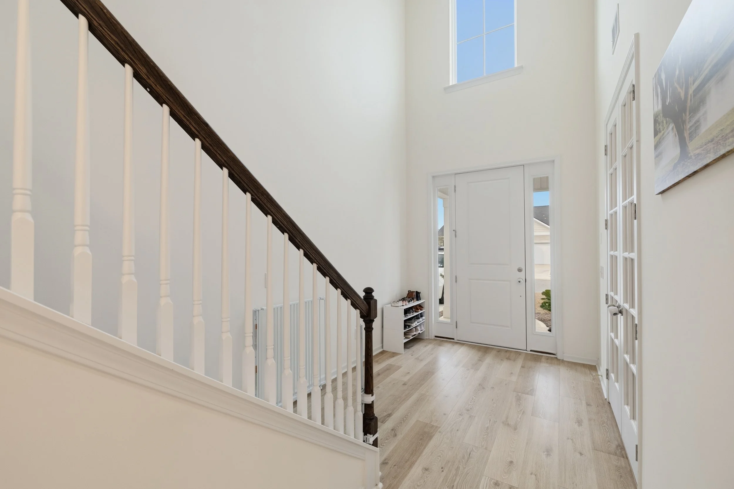 Entryway of a house with a white front door, two tall vertical windows on either side, a staircase with a dark wooden handrail and white spindles, hardwood flooring, a small shoe rack, and a painting of a tree on the wall.