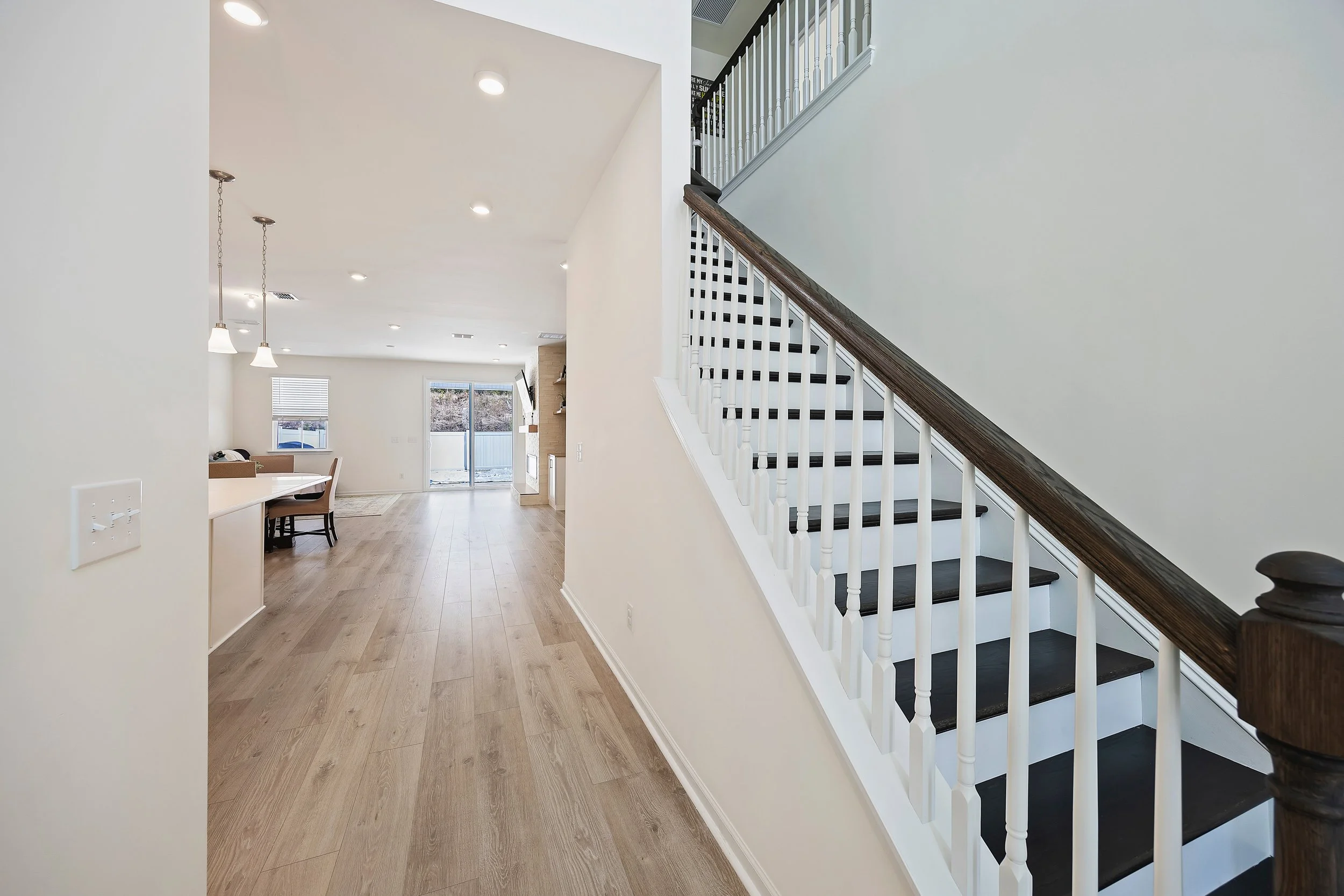 Interior of a modern home with a staircase featuring dark wood steps and white spindles, leading to the upper floor. The area is bright with recessed lighting, hardwood floors, and an open layout that includes a dining area with a hanging light fixtu
