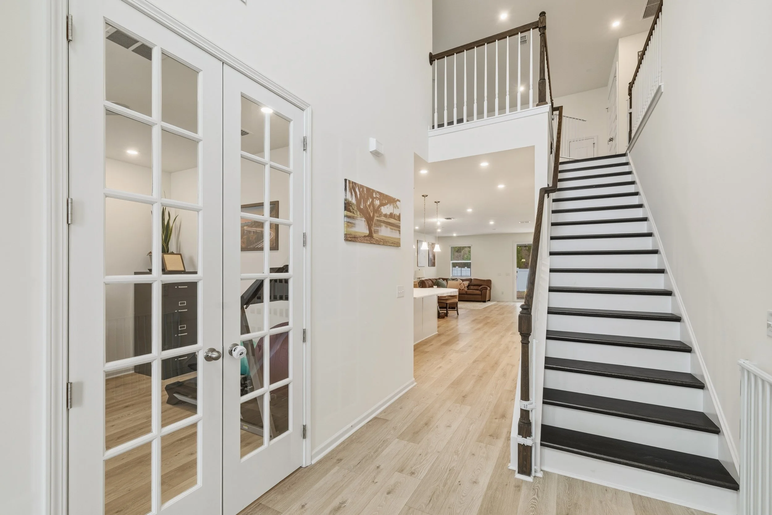 Interior view of a modern house with a staircase, glass-paneled double doors, and an open living room with a brown sofa and windows.