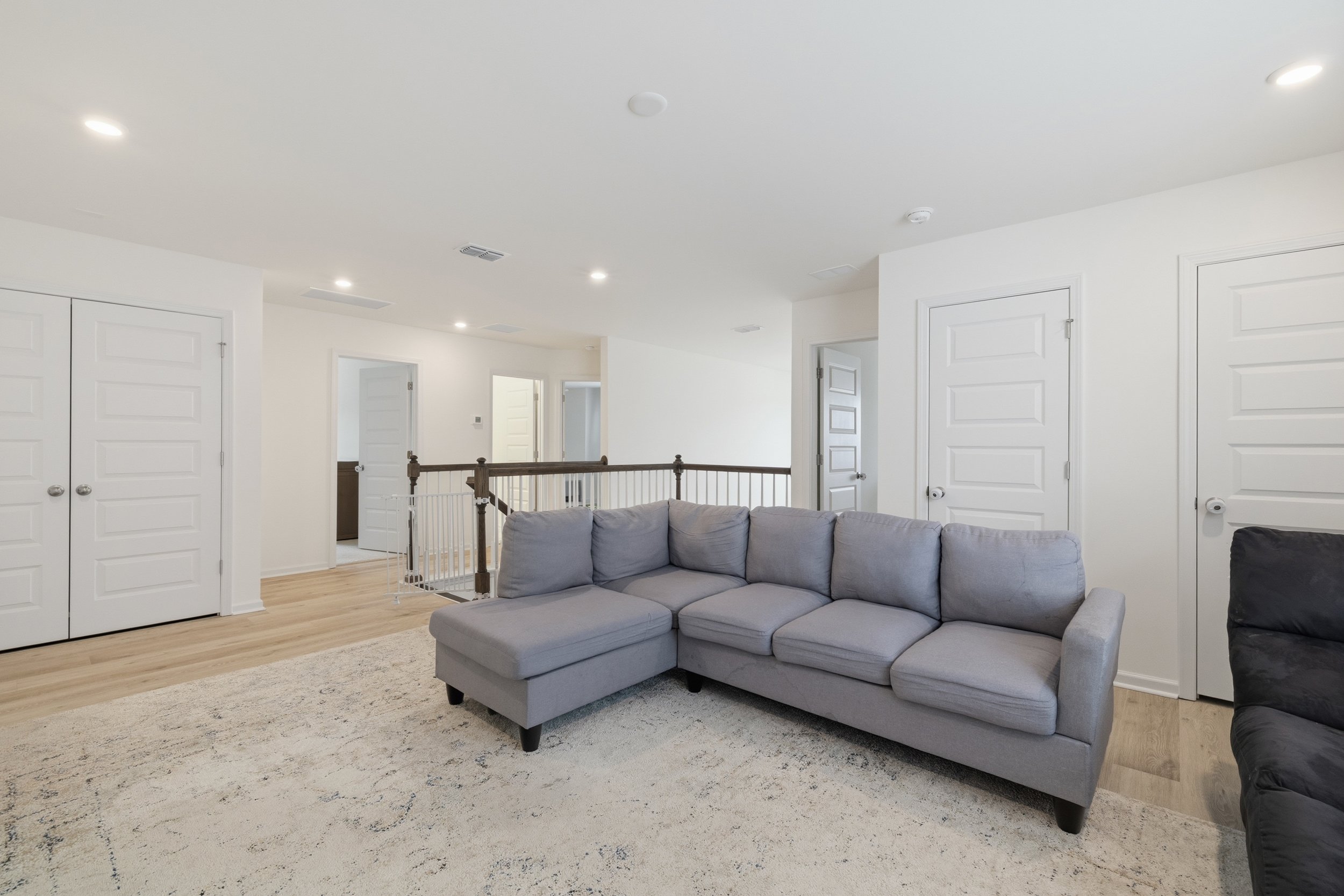 Living room with gray sectional couch, white walls, light wood flooring, and multiple closed white doors.