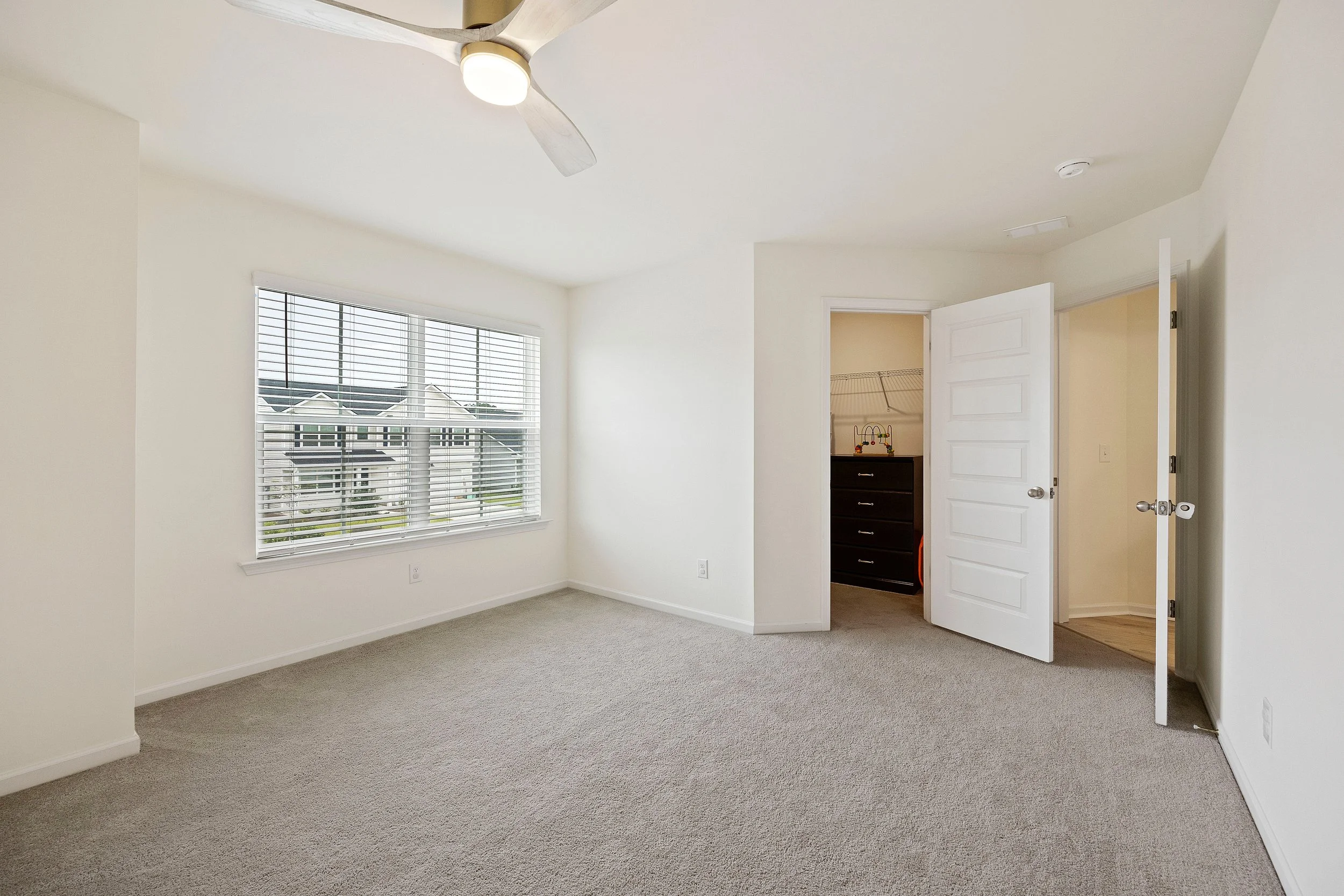 Empty bedroom with a large window, beige carpet, white walls, a ceiling fan, and an open closet with a dark dresser inside.
