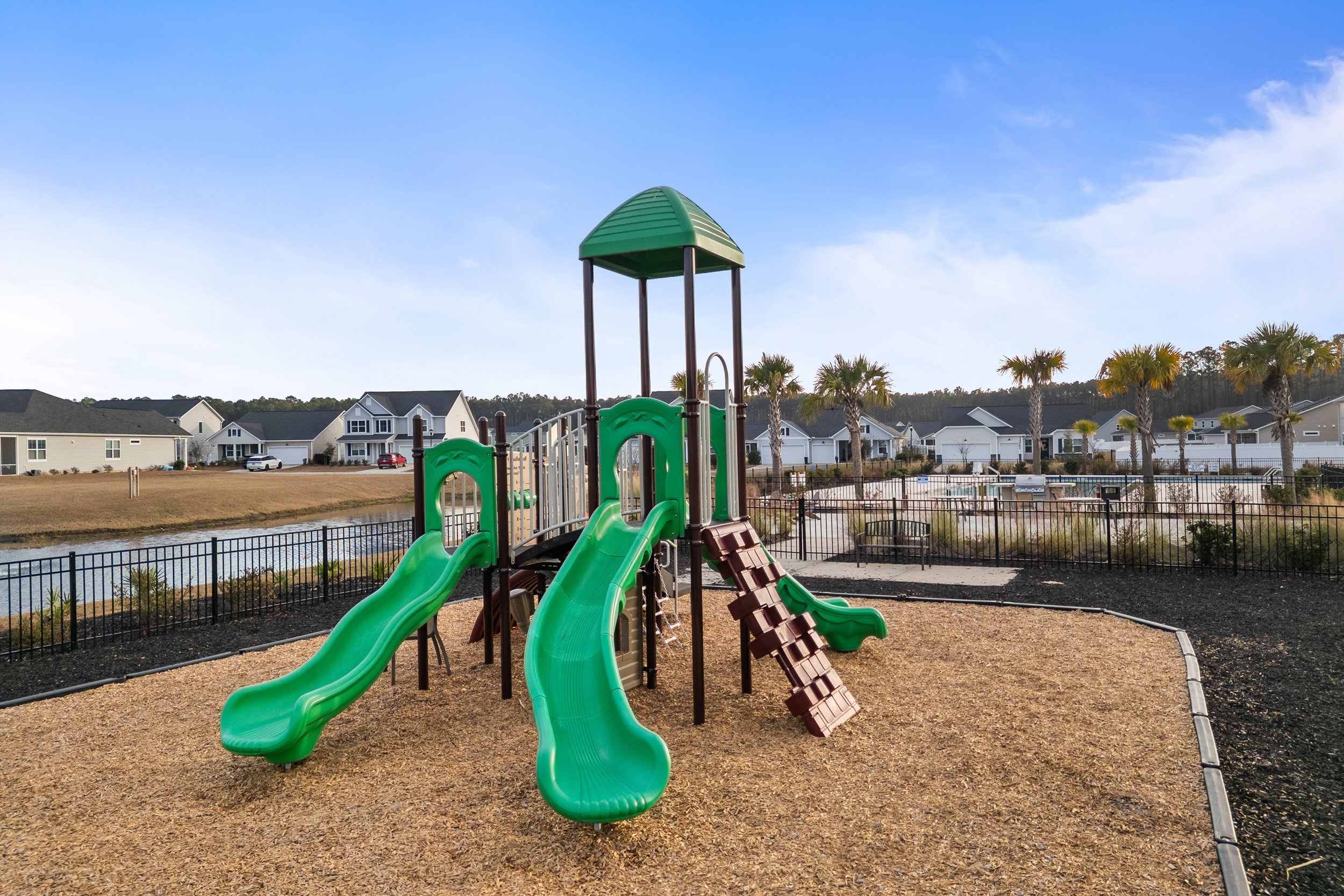 Playground with green slides, brown climbing steps, and a small covered platform in a fenced yard near a pond, with houses and palm trees in the background under a partly cloudy sky.