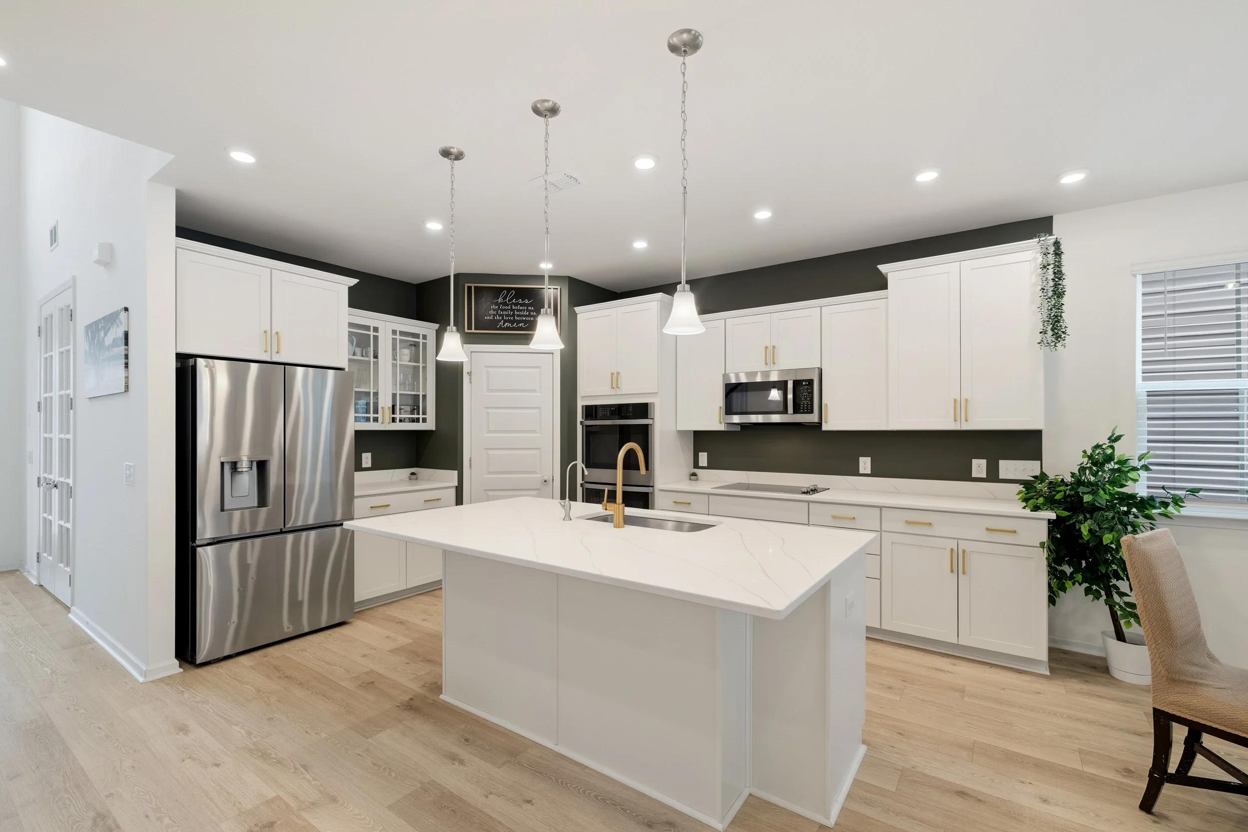 Modern kitchen with white cabinets, a large island with a sink, stainless steel refrigerator, microwave, oven, and a dark accent wall with a framed quote and a hanging plant, illuminated by ceiling and pendant lights.