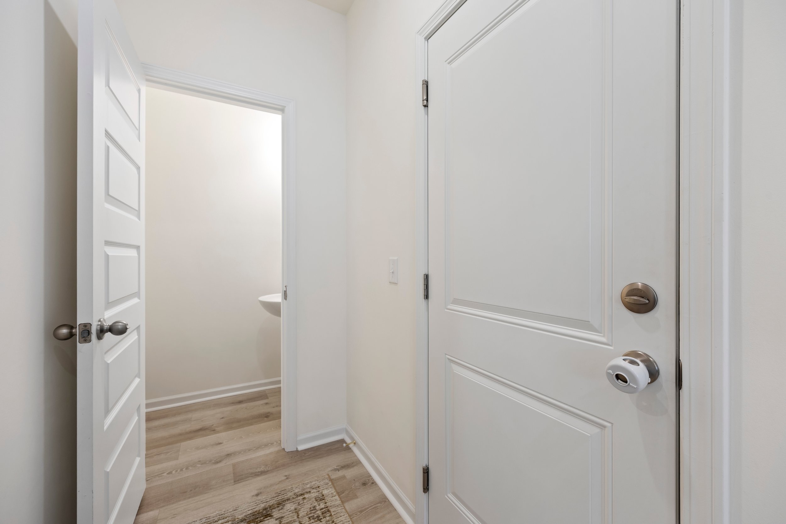 View of a hallway with a closed white door on right and an open door revealing a small bathroom with a wall-mounted sink, hardwood flooring, and white walls.