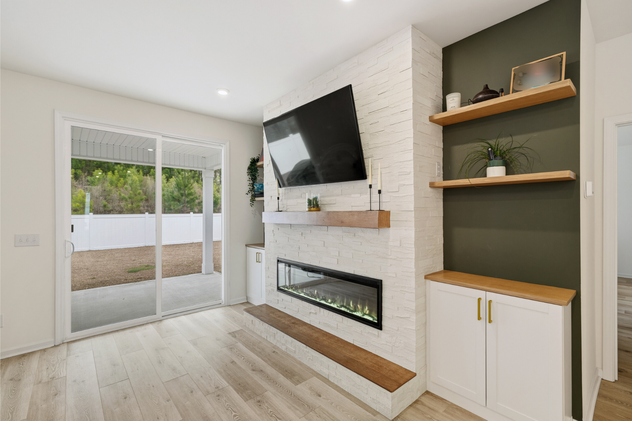 Living room with a white brick fireplace, mounted TV, sliding glass door to outside, and dark green accent wall with wooden shelves holding decor and plants.